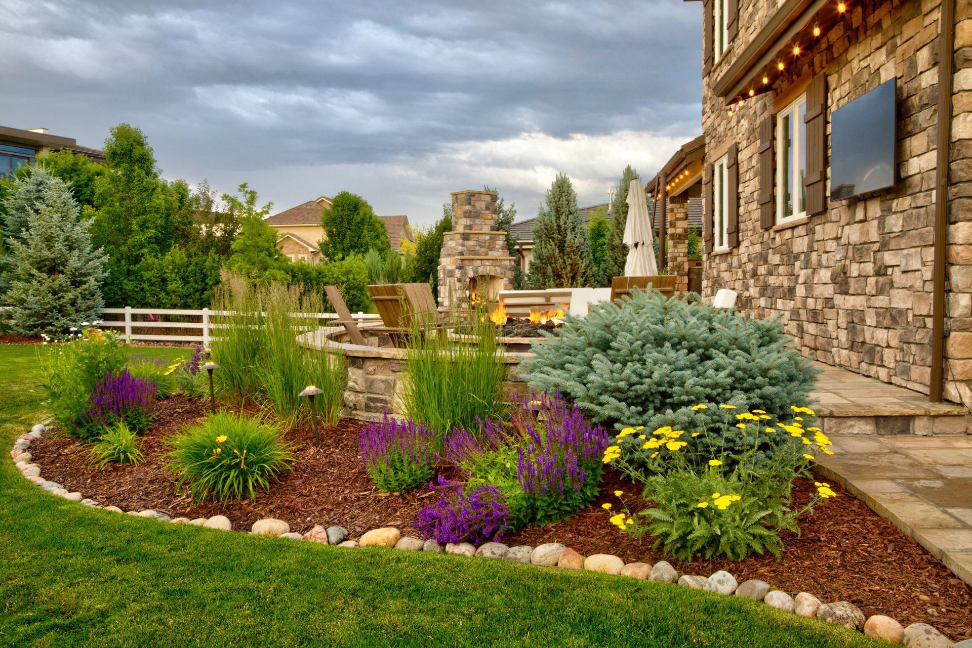 A lush garden with purple and yellow flowers, a blue spruce, and a stone patio with a fireplace against a house exterior.