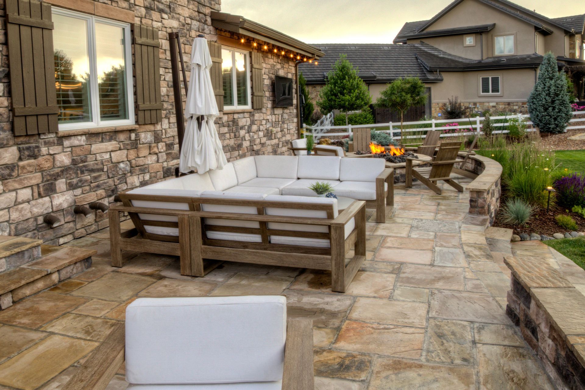 A stone patio with a modular outdoor sofa, a fire pit, and lounge chairs next to a rustic home at dusk.