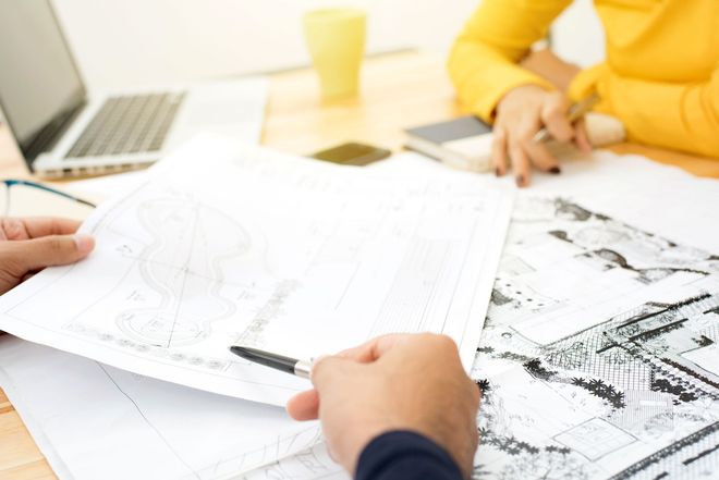 Two people working together at a desk, reviewing technical blueprints and drawings with a pen.