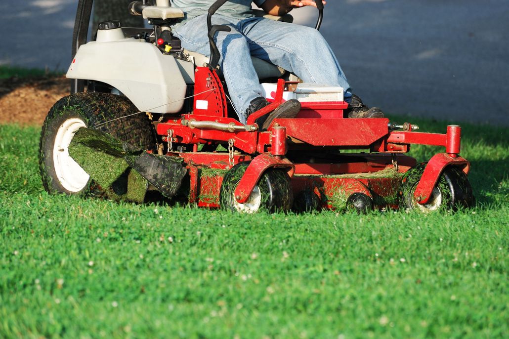 A person sits on a red zero-turn lawn mower, cutting green grass on a sunny day.