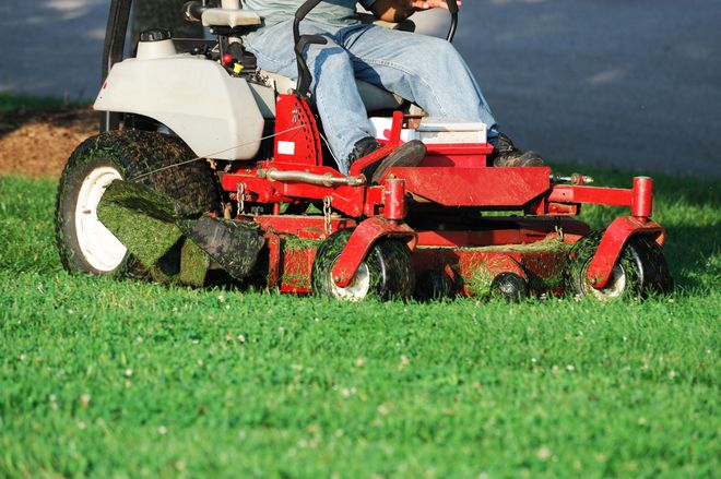 A person sits on a red zero-turn lawn mower, cutting green grass on a sunny day.