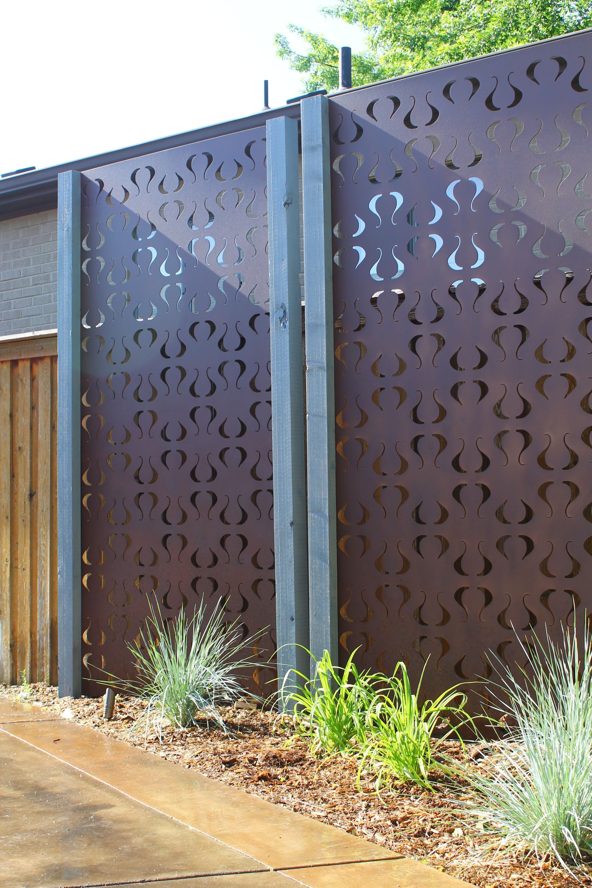 A metal privacy screen with a patterned cut-out design, mounted on dark pillars in a landscaped garden bed.