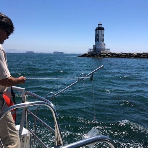 Person on a boat points a pole toward a lighthouse on a rocky breakwater under a clear blue sky.