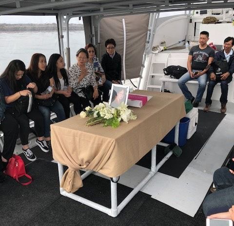 People seated around a coffin with flowers on a boat deck during a memorial service