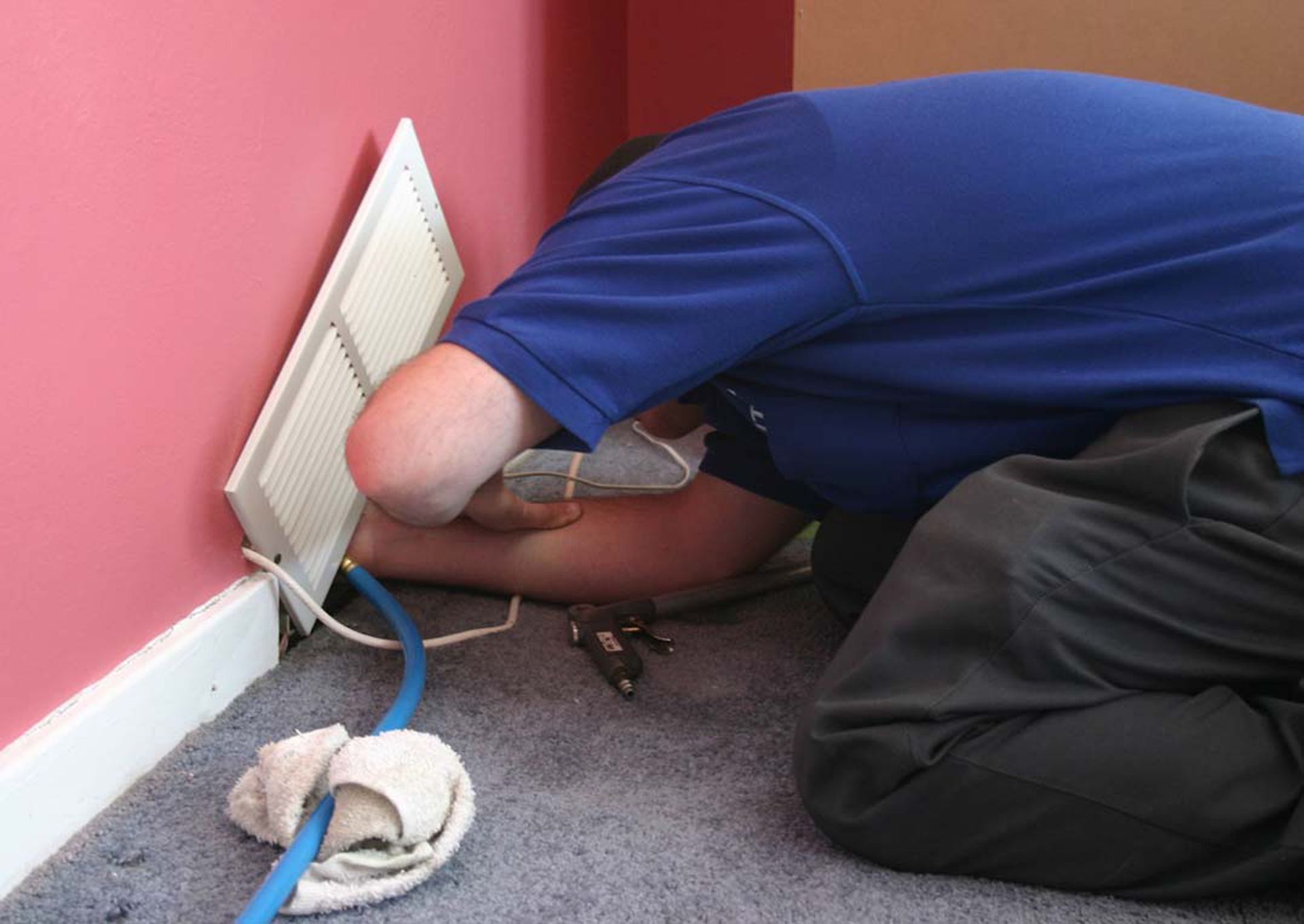 Person kneeling beside a wall vent, reaching into an opening with tools and cables on the carpet.