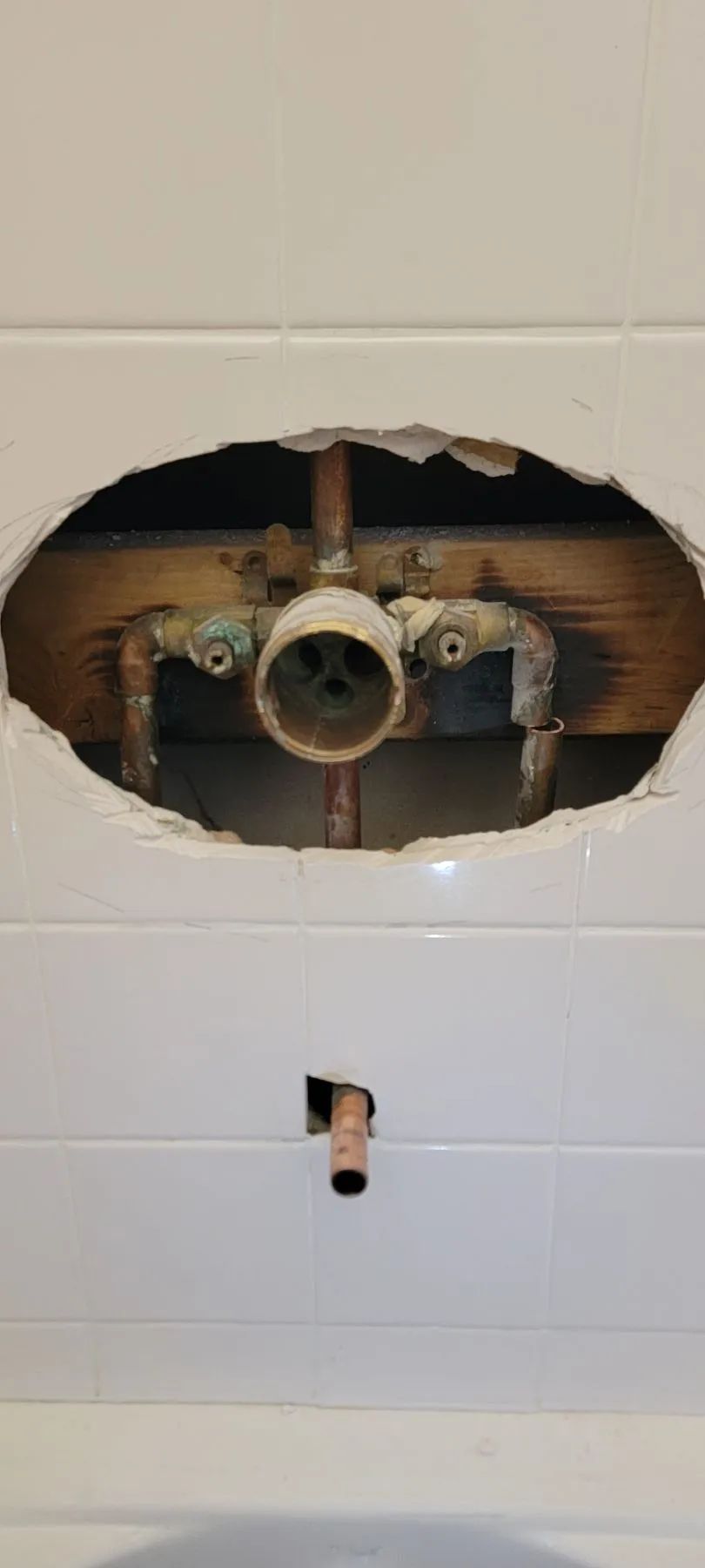 Bathroom wall with a large hole exposing plumbing pipes and wiring behind white tiles.