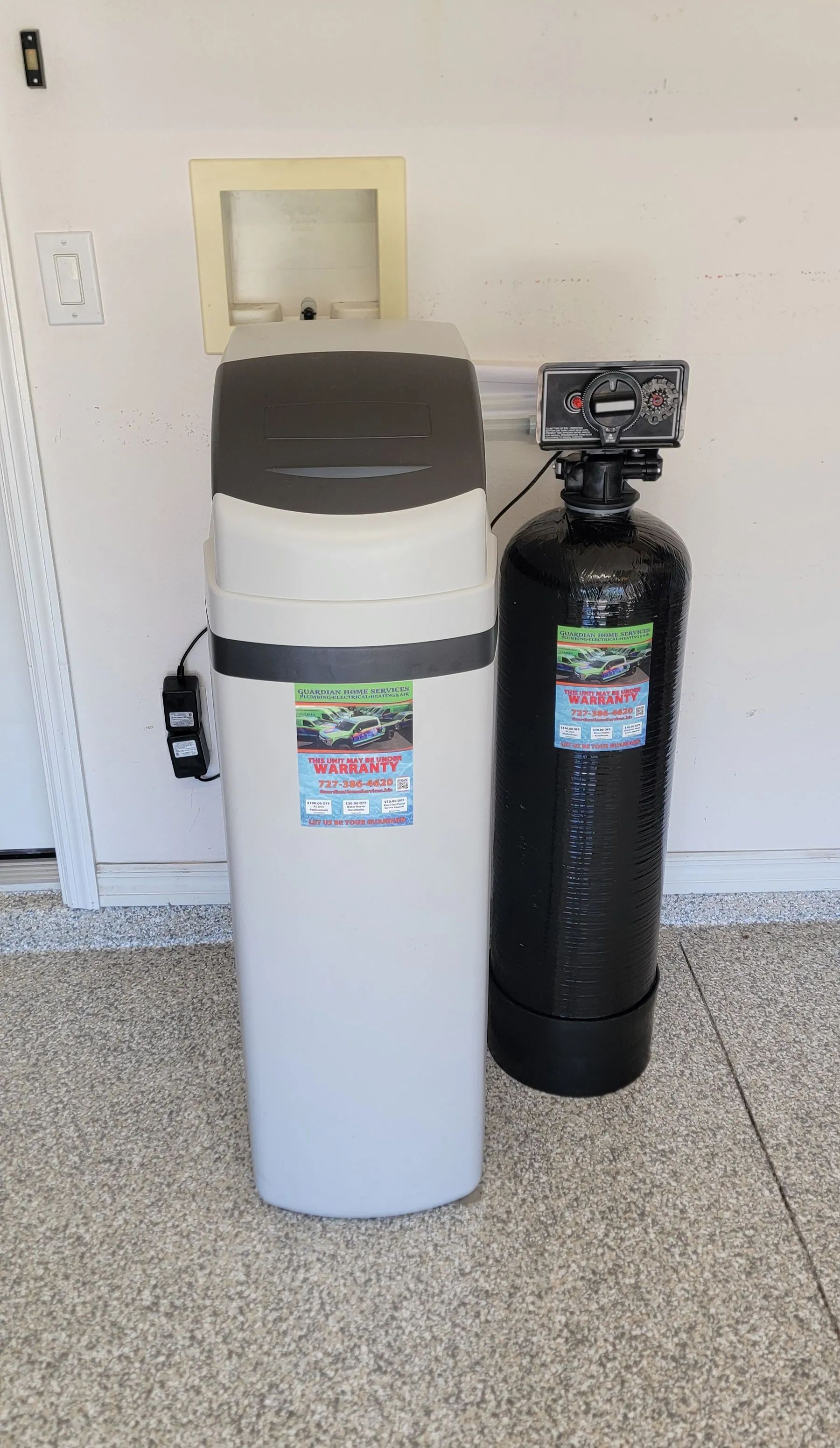 Two water softener tanks against a white wall, one light gray and one black, in a garage-like room.