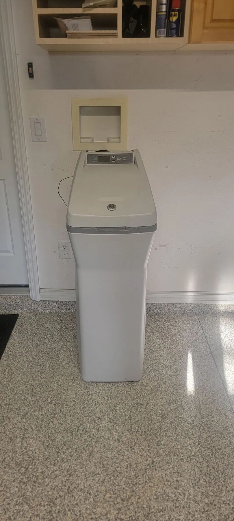 White top-loading washing machine in a laundry room, with cabinets above and speckled flooring.