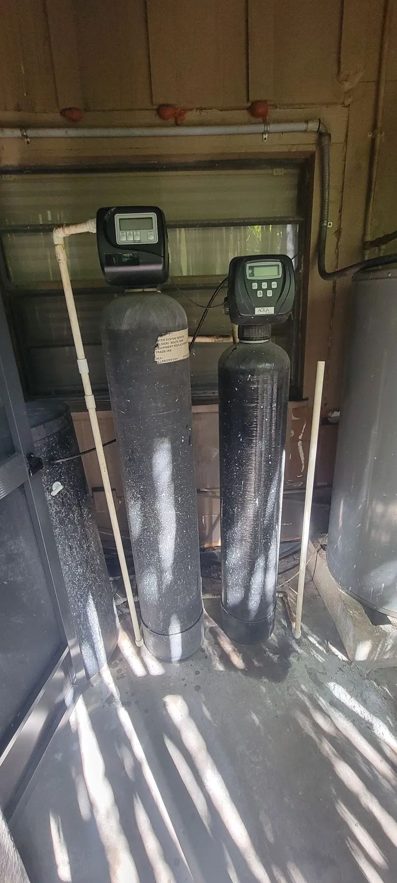 Two black water softener tanks in a utility room, lit by sunlight through slats.