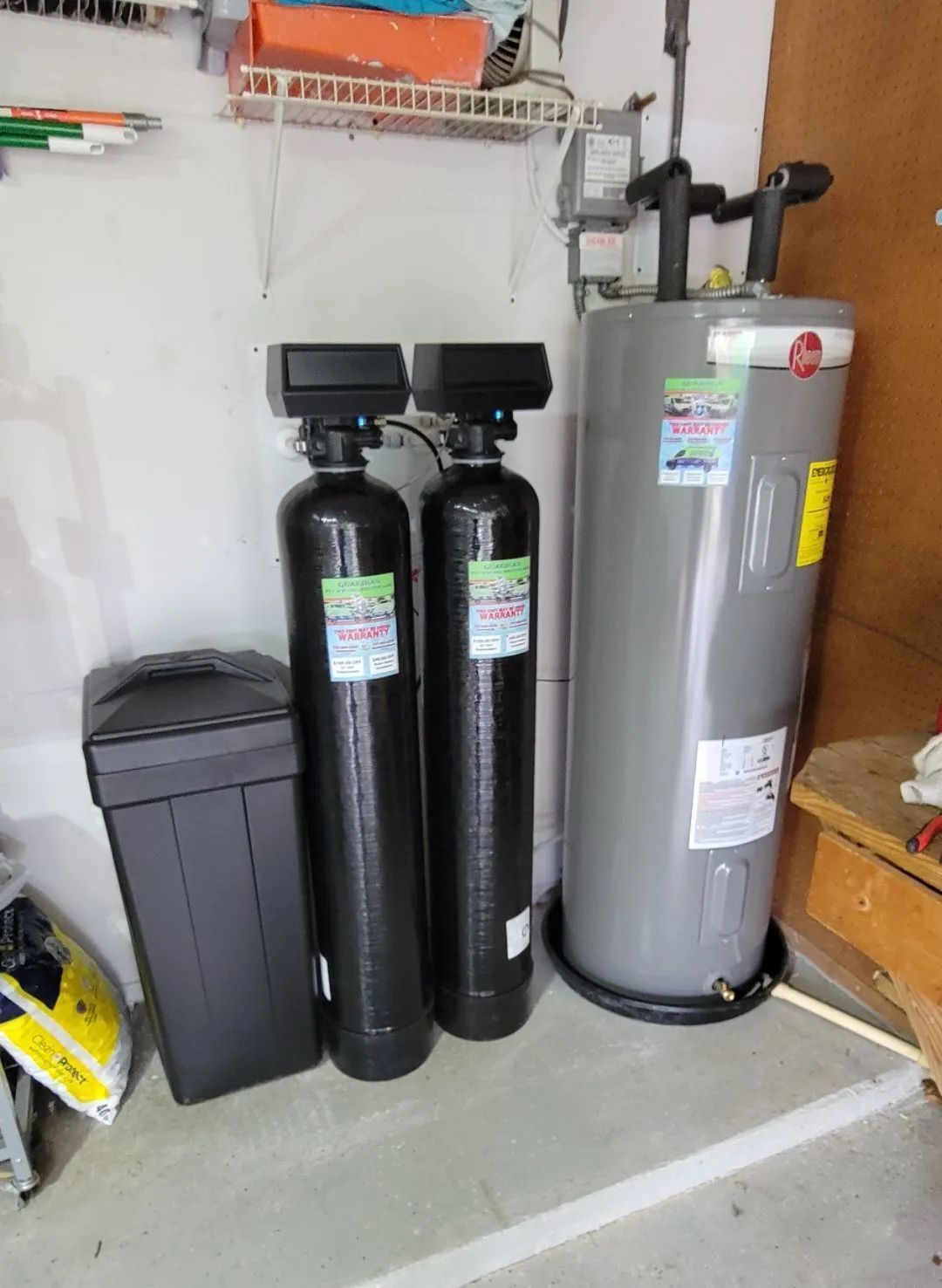 Water softener equipment and a tall gray tank in a garage beside a black trash bin.