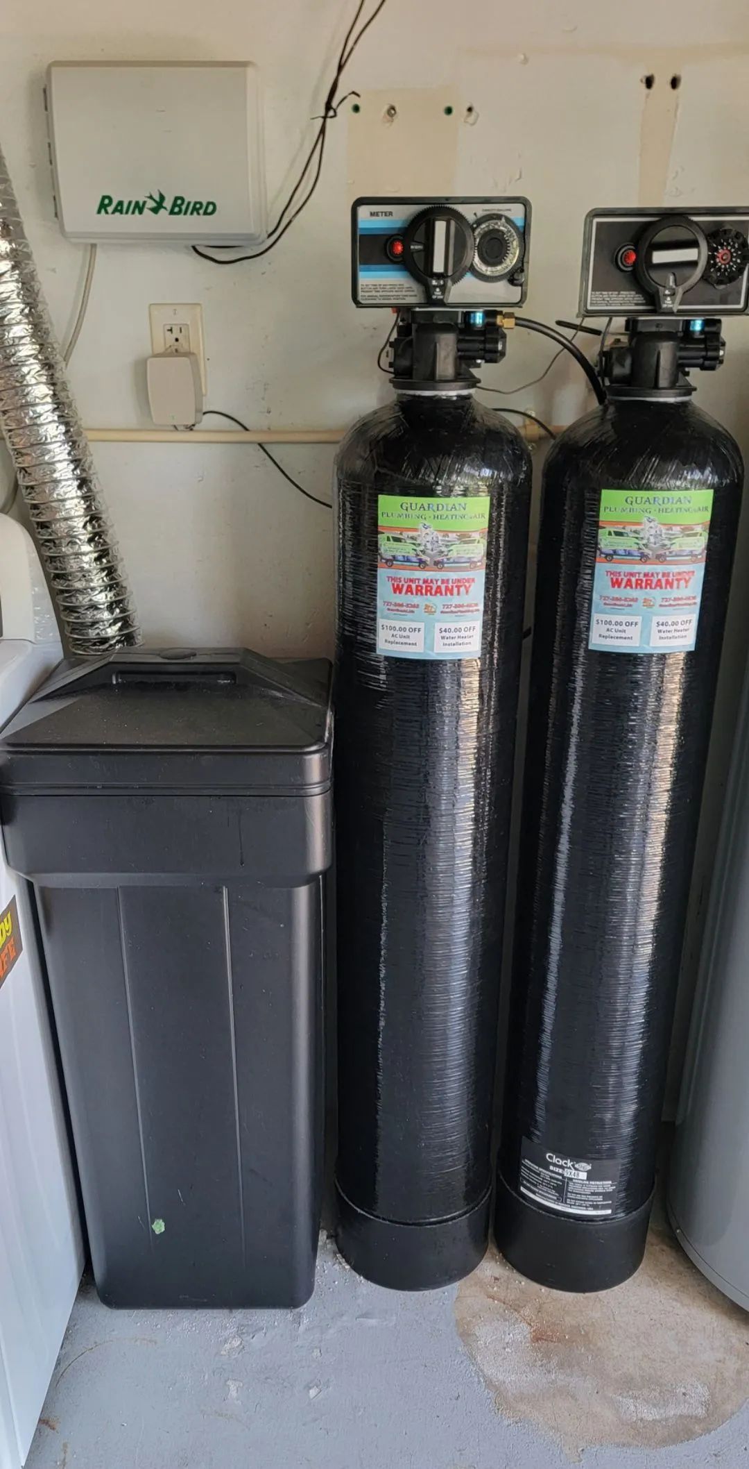 Black water softener tanks beside a trash bin in a garage or utility room