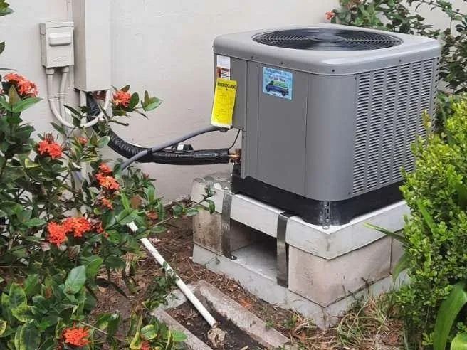 Outdoor air conditioner unit beside bushes and flowers on a concrete pad