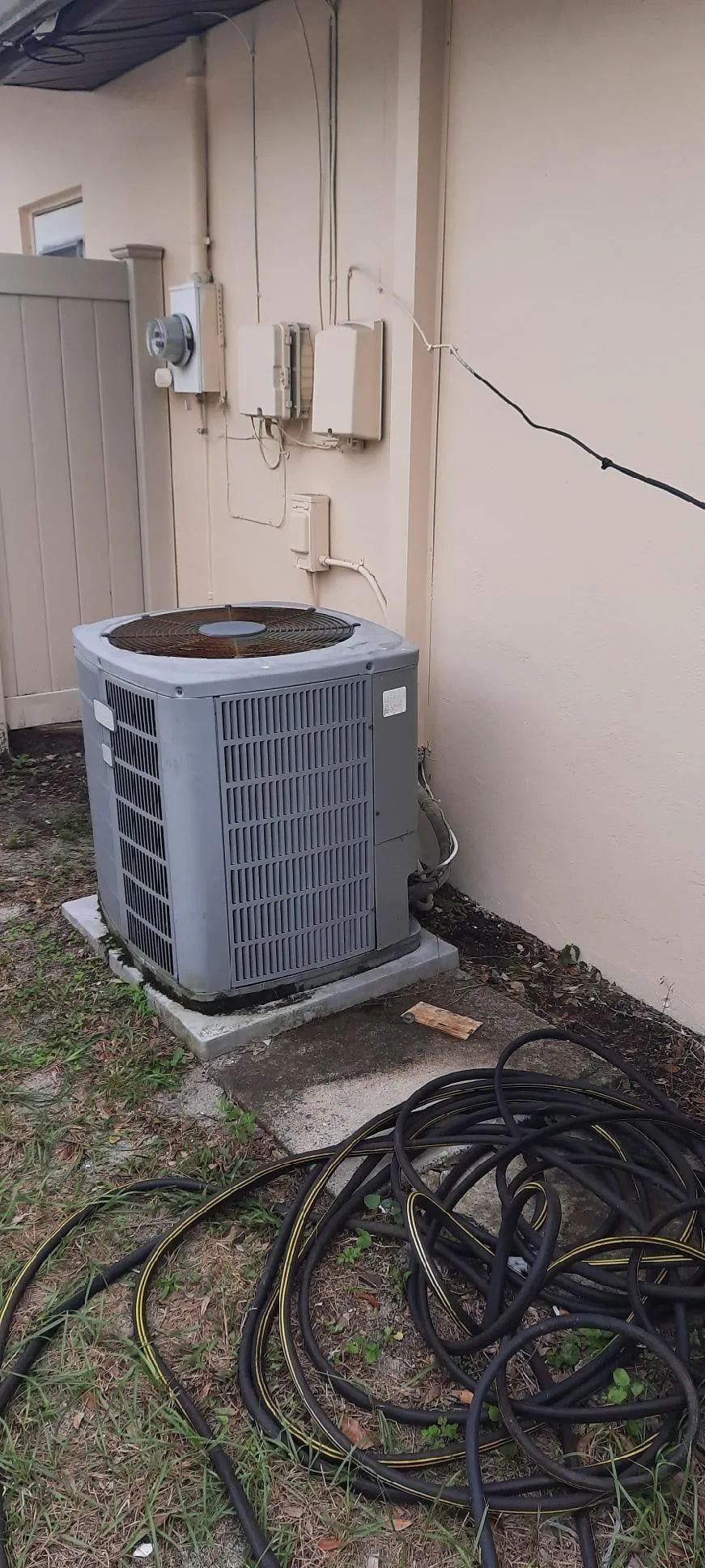 Outdoor HVAC unit beside a beige wall, with tangled black cables on the ground.