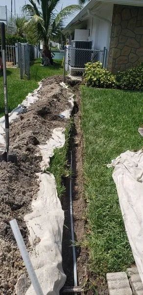Long trench dug beside a house, with exposed pipes and piled dirt along a grassy yard.