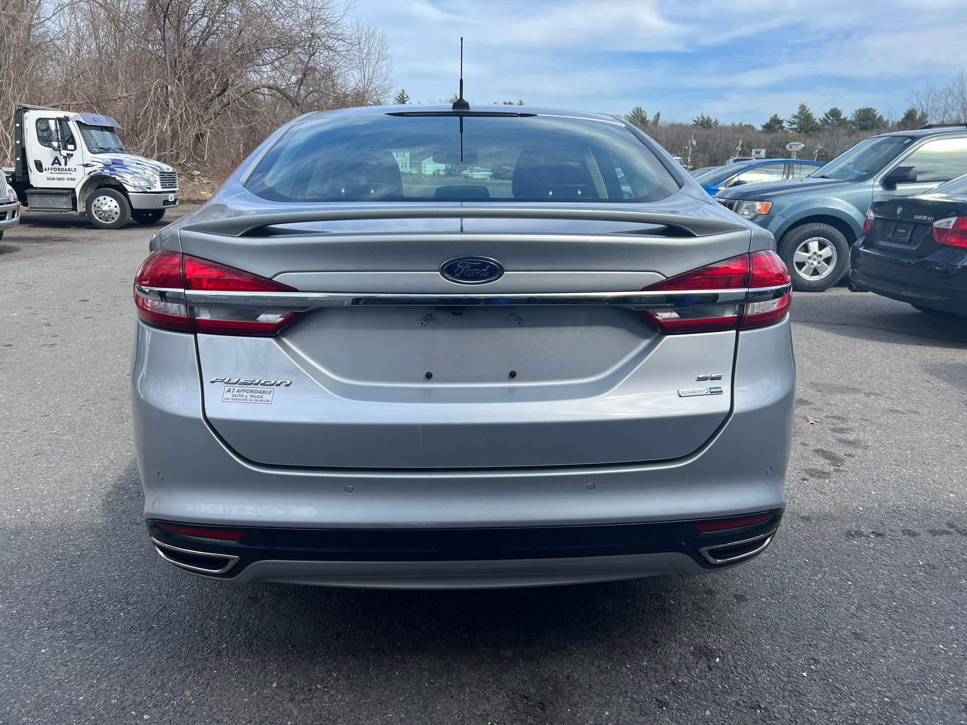 A silver ford fusion is parked in a parking lot next to a tow truck.