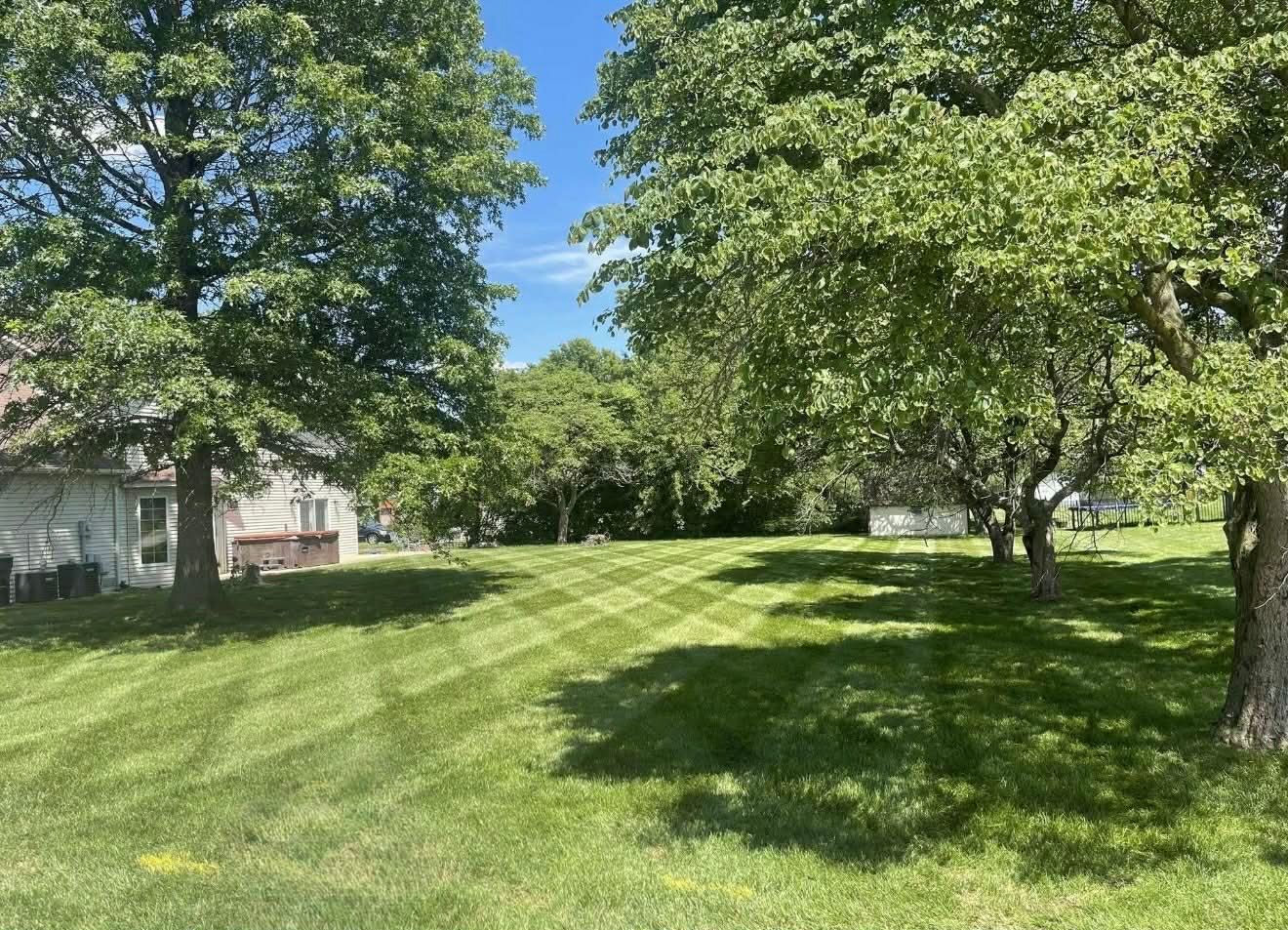 A lush green lawn with crisp, diamond-patterned mower stripes, flanked by large leafy trees under a bright blue sky.