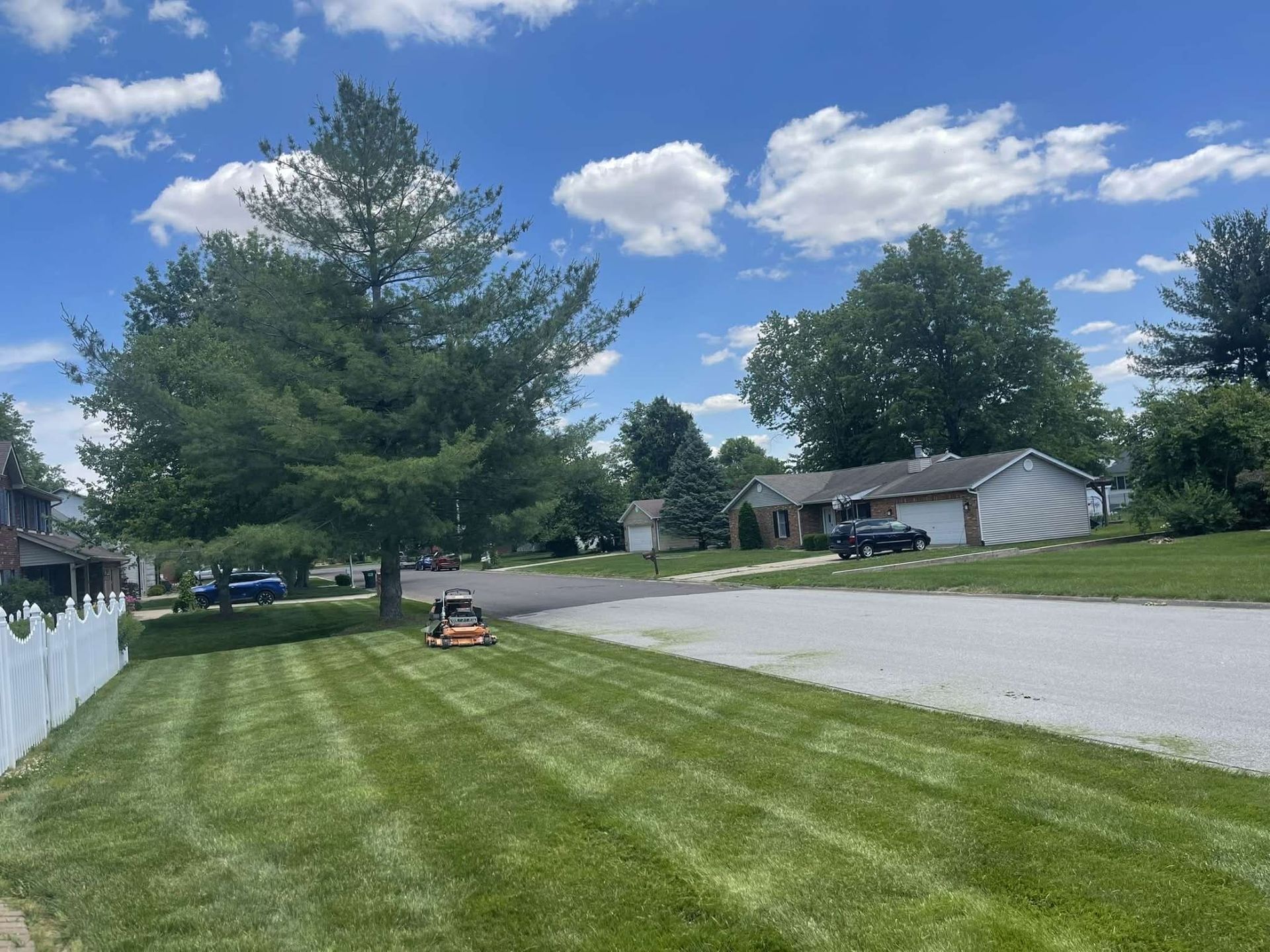 A person on a riding lawnmower cuts grass in a suburban yard with a house and trees under a blue sky with clouds.