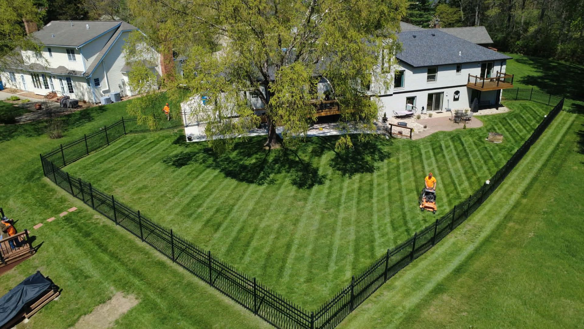 A drone view of a landscaped suburban backyard with neatly mown lawn stripes enclosed by a black metal fence.