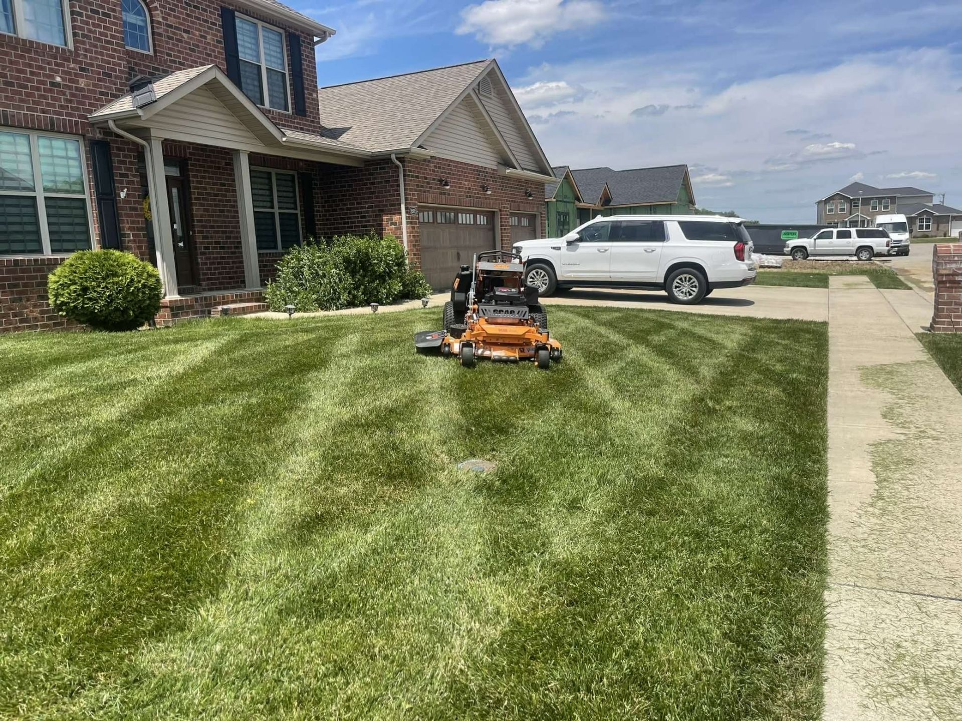 A bright orange lawn mower sits in the center of a freshly mowed front lawn next to a brick house and a white SUV.