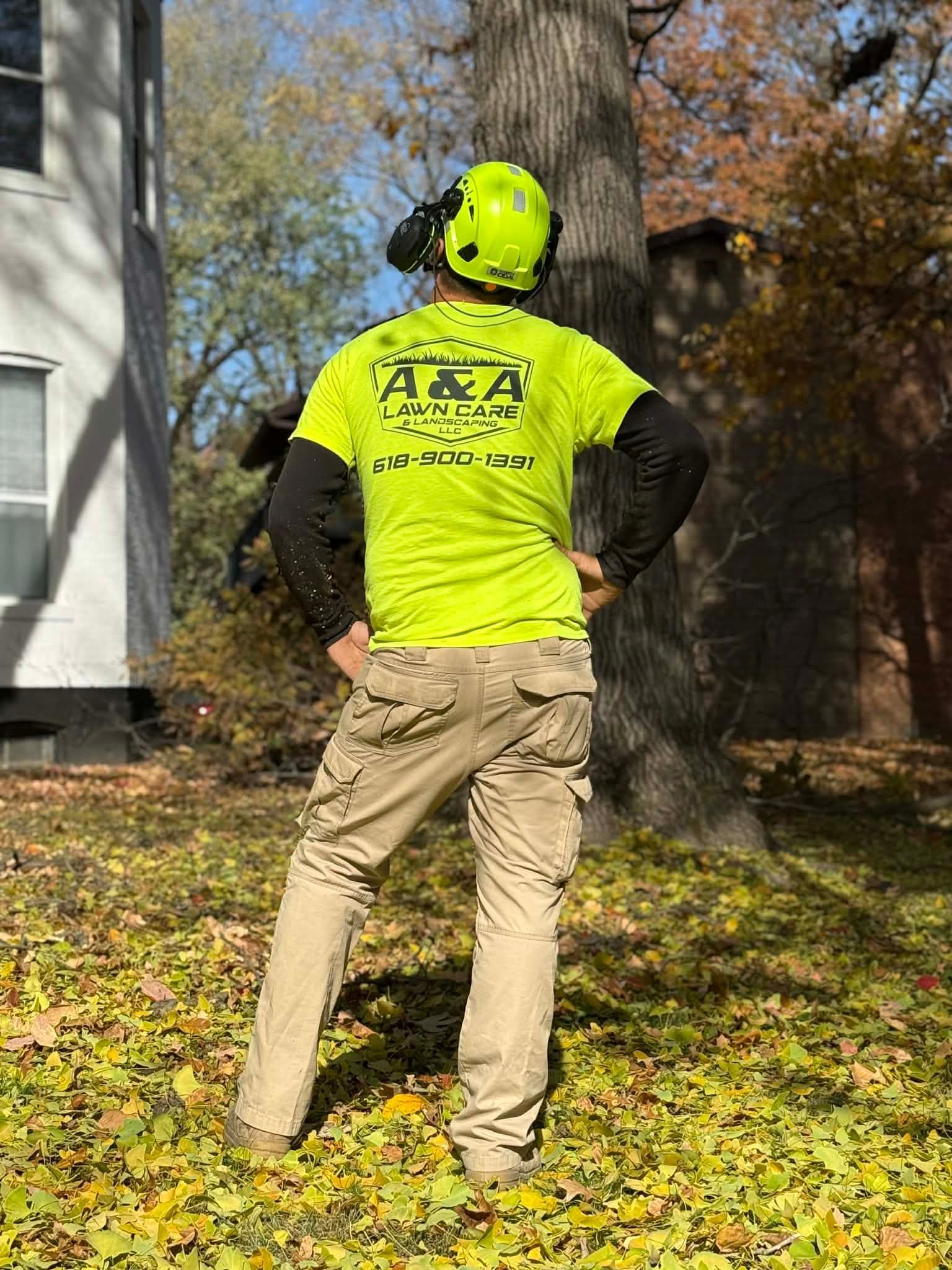 A worker in a bright yellow shirt and safety helmet standing in a yard covered in autumn leaves, facing a large tree.