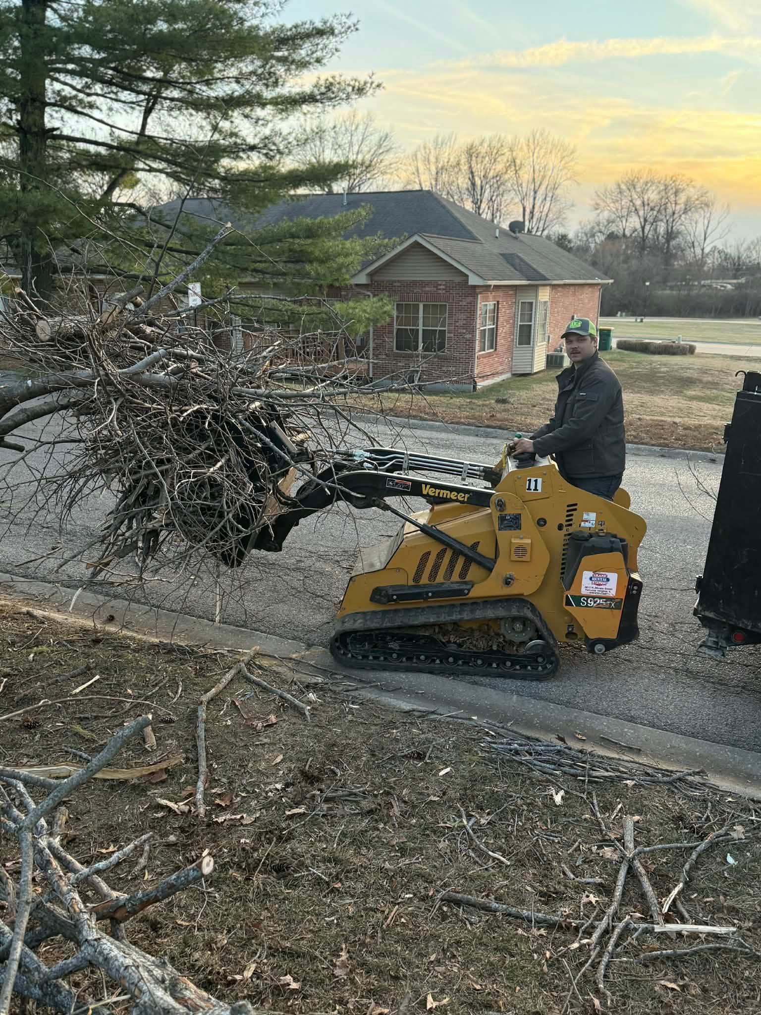 A person operates a yellow tracked skid steer loader to clear brush and tree limbs from a residential street.