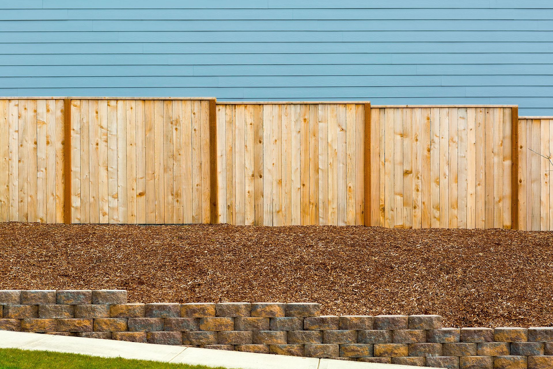 Wooden fence on top of brown gravel with tiered stone wall in front. Blue sky background.