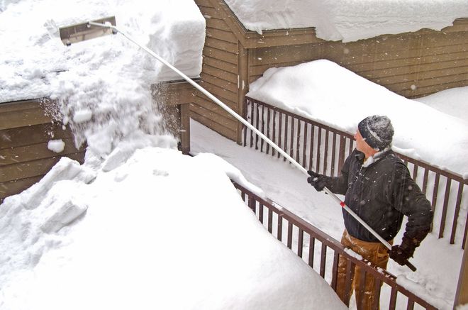 Man removing snow from a roof with a long-handled snow rake. Snow covers the deck and walls.