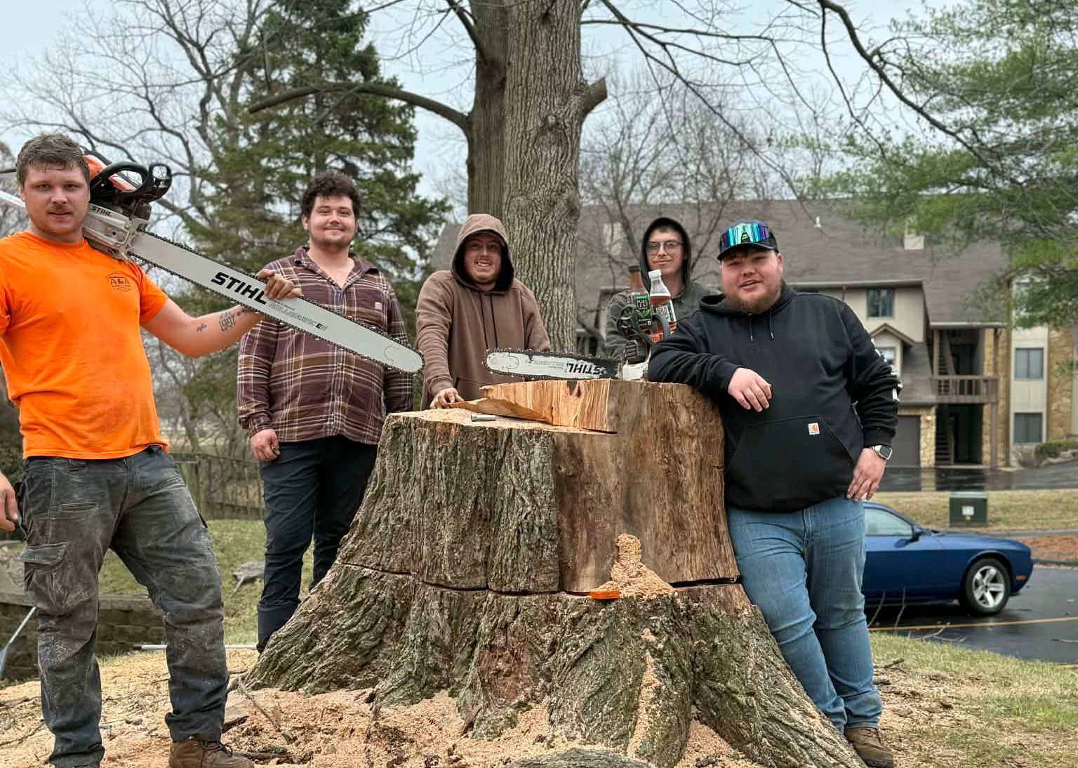 Five people pose outdoors next to a large tree stump with a chainsaw, with a car and buildings in the background.