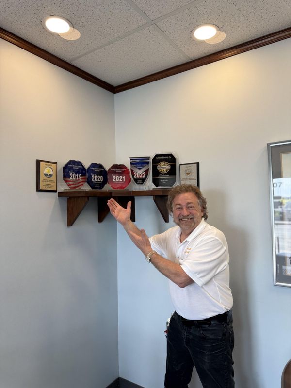 A smiling person in a white polo shirt gestures toward a wooden corner shelf displaying several award plaques.