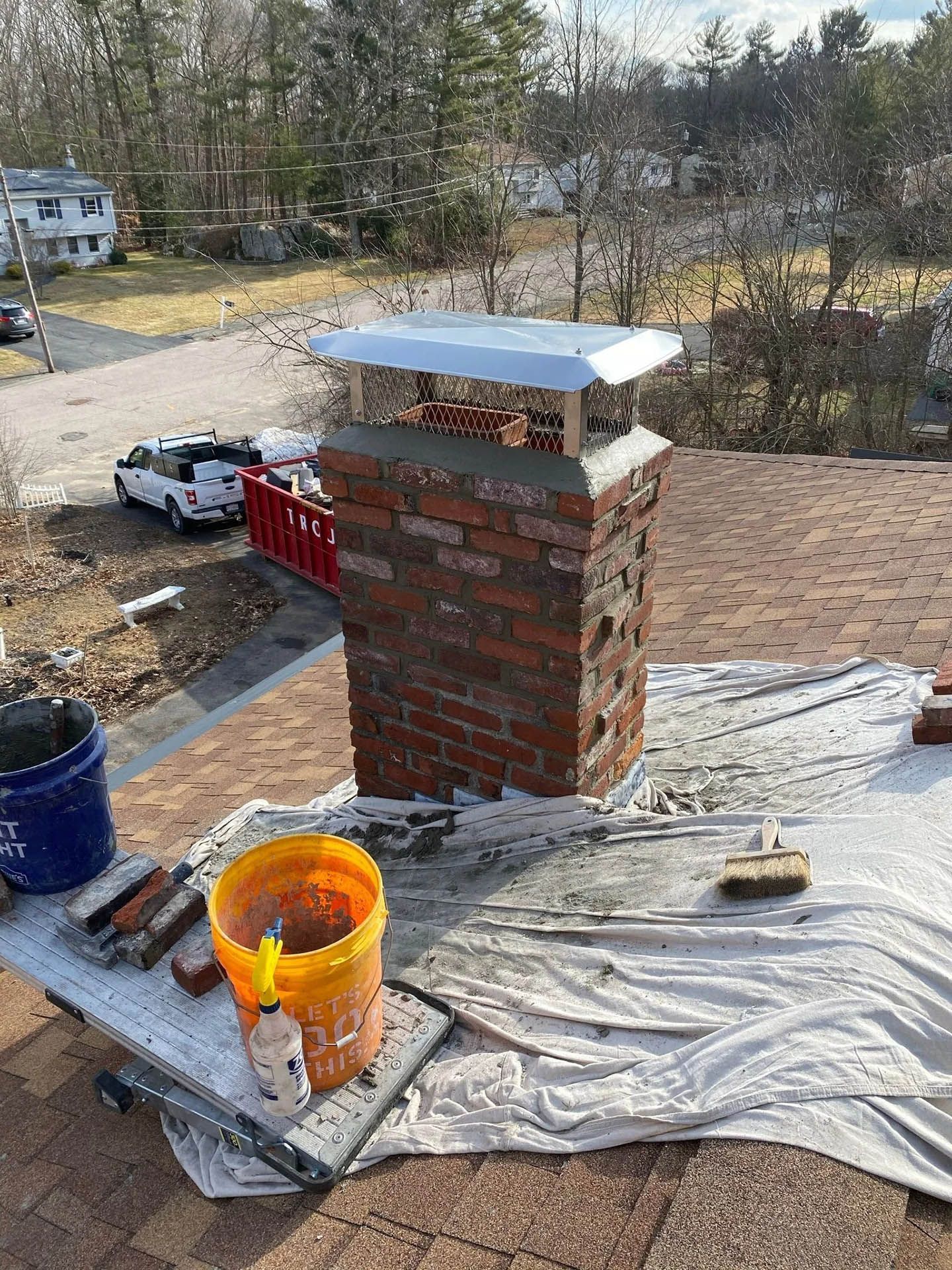 A brick chimney on a residential roof, featuring a new metal cap, with construction tools and buckets nearby.
