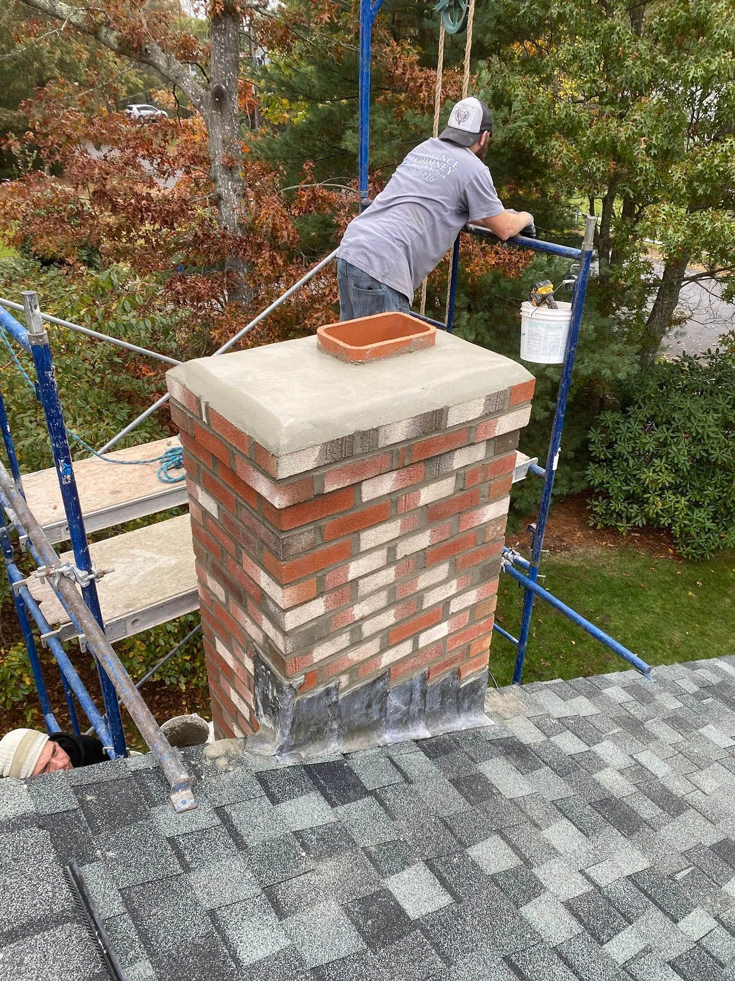 A worker repairs a brick chimney on a roof, standing on blue scaffolding with a concrete cap and red flue tile.