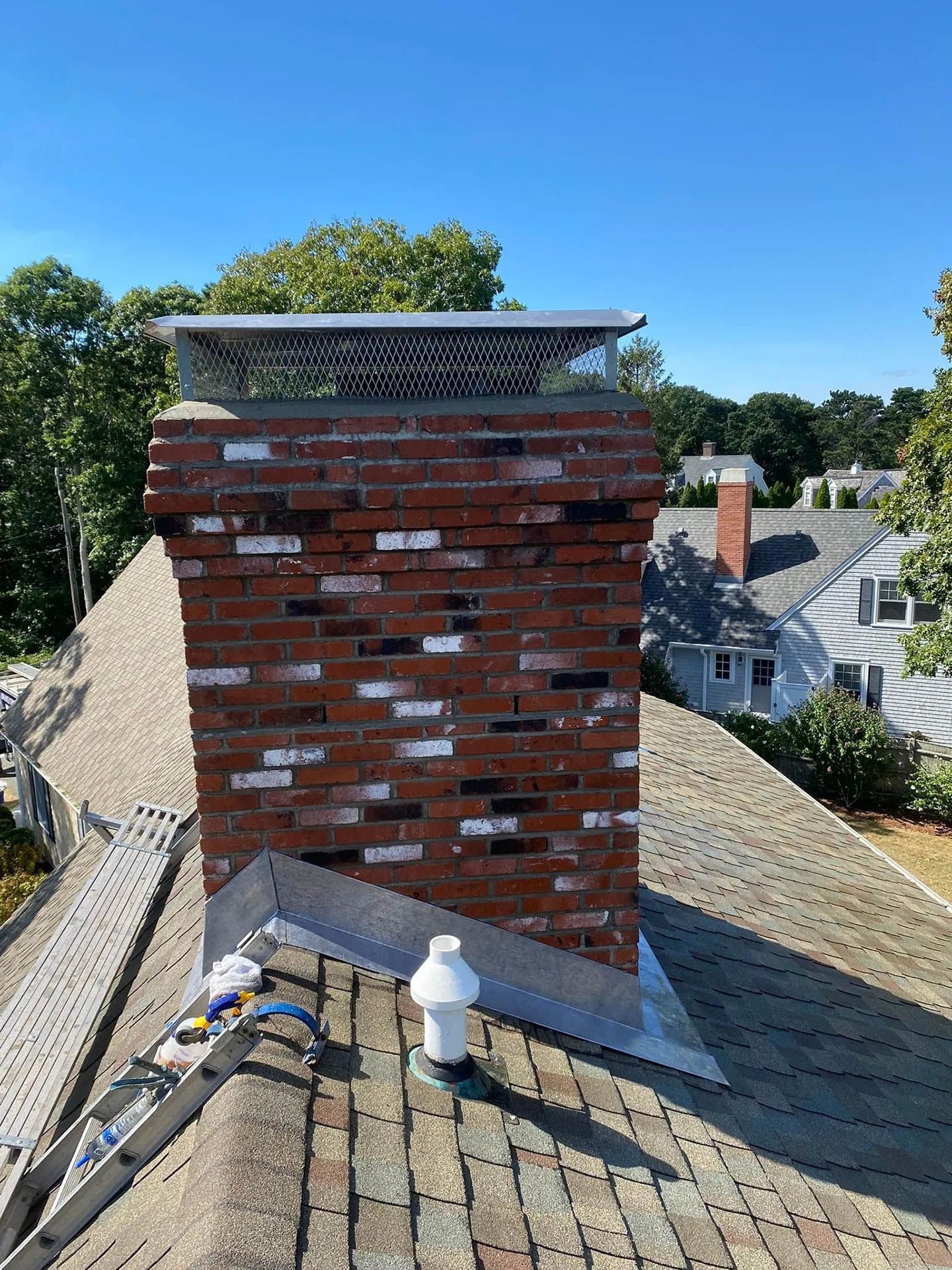 A brick chimney on a shingled roof with a metal rain cap and flashing, seen under a clear blue sky.