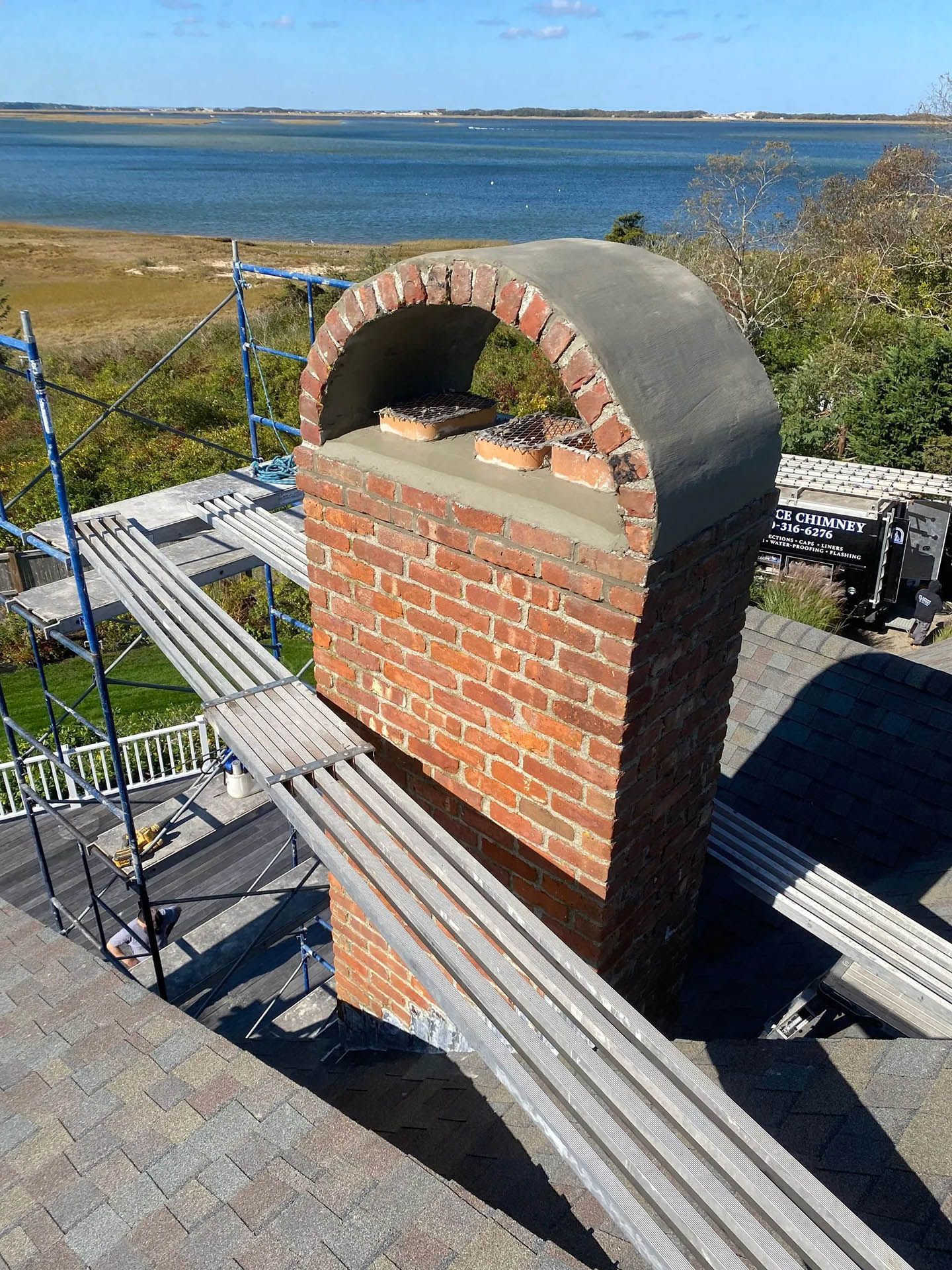 A tall brick chimney with a decorative arched brick cap stands on a rooftop, surrounded by scaffolding overlooking a bay.