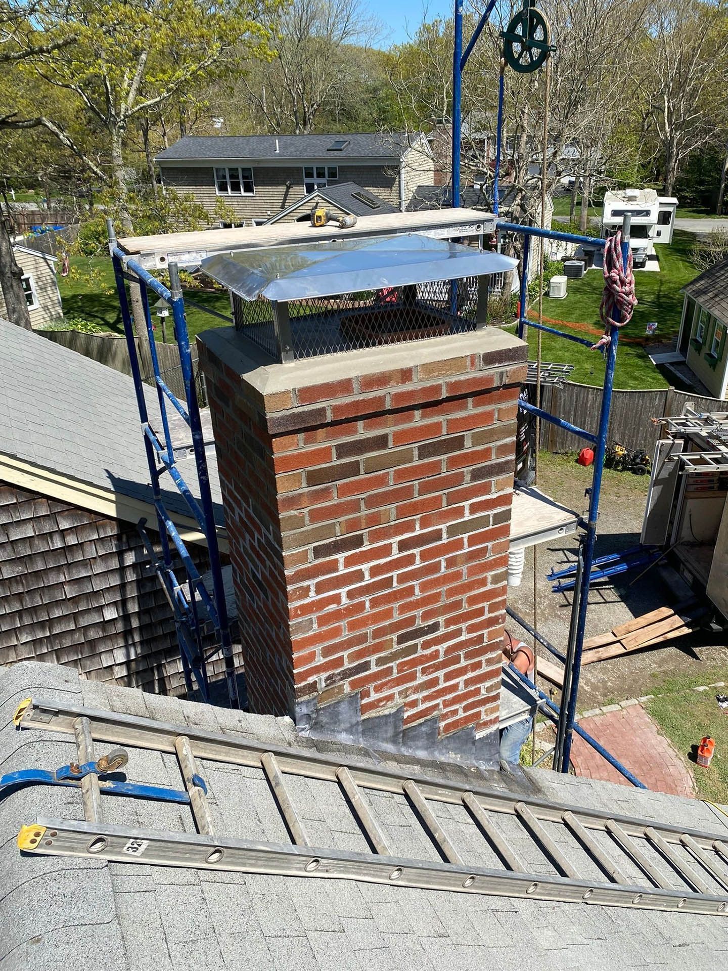 A brick chimney on a residential roof, surrounded by blue scaffolding, with a metal chimney cap installed at the top.
