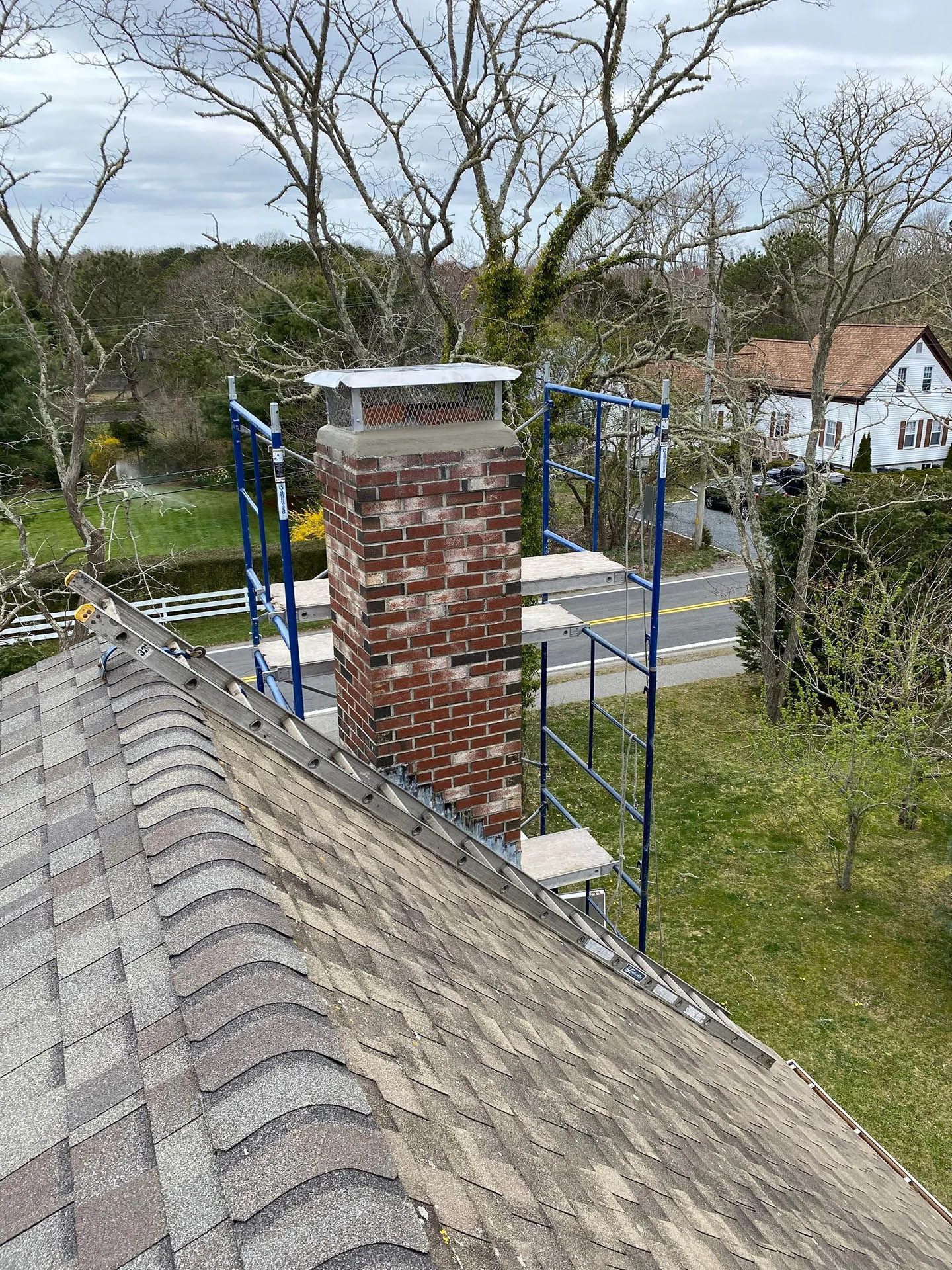 A red brick chimney with a metal cap stands on a shingled roof, surrounded by blue scaffolding with a road in the distance.