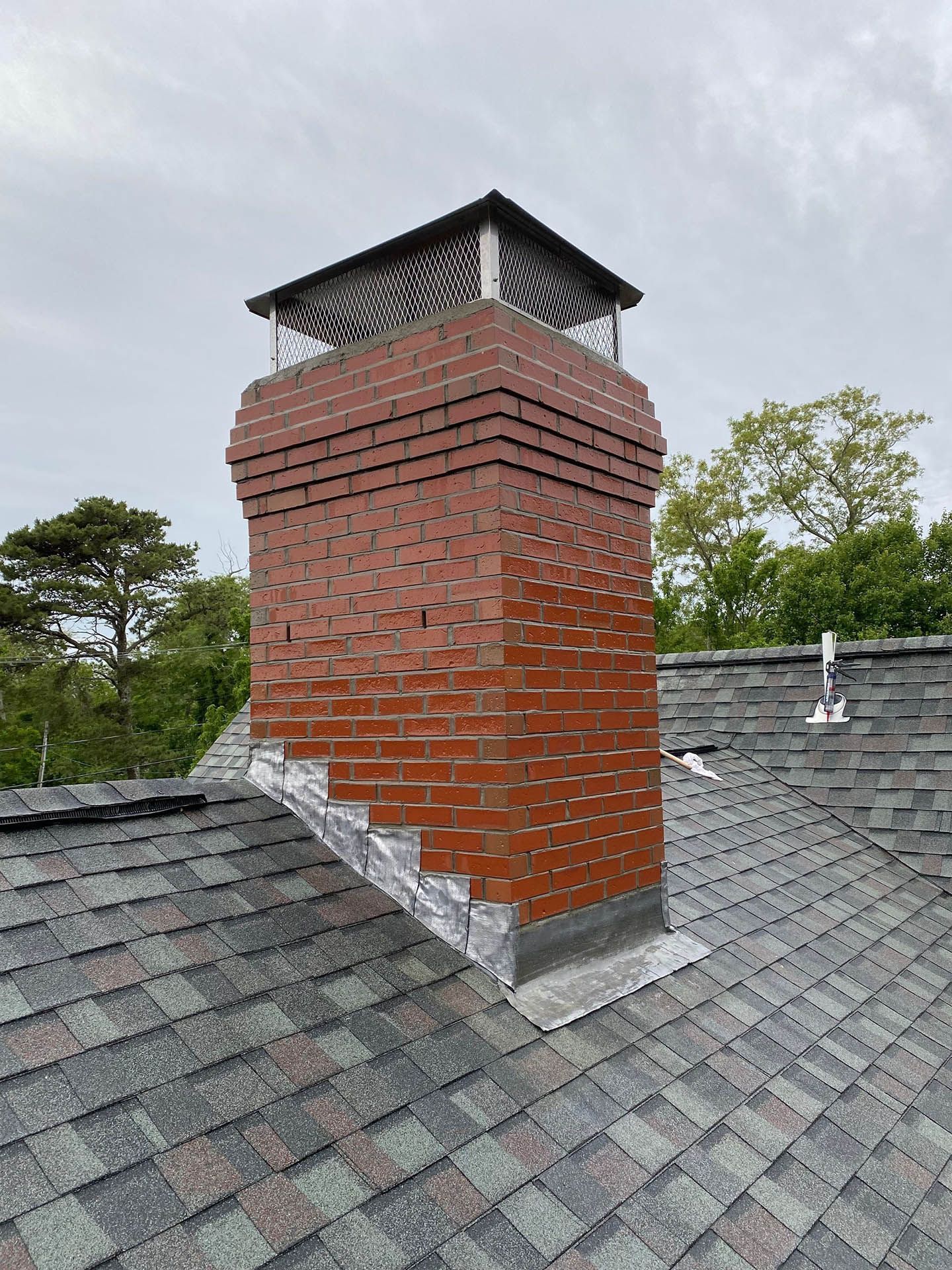 A red brick chimney with a metal cap and flashing, situated on a dark grey shingled roof under a cloudy sky.