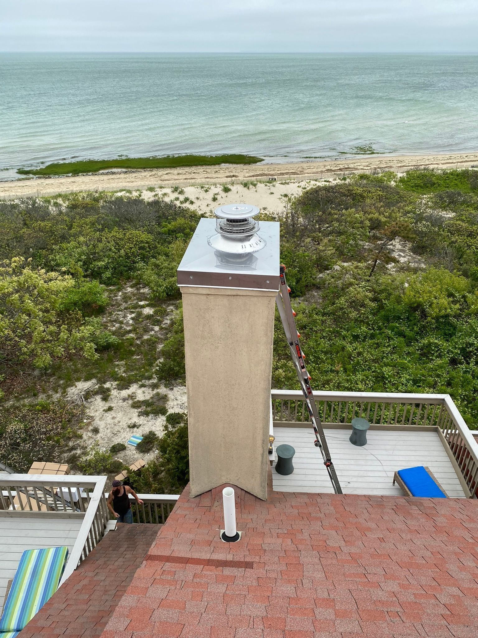 A tall stucco chimney with a metal cap sits on a reddish roof overlooking a beach and the ocean, with a ladder leaning by it.