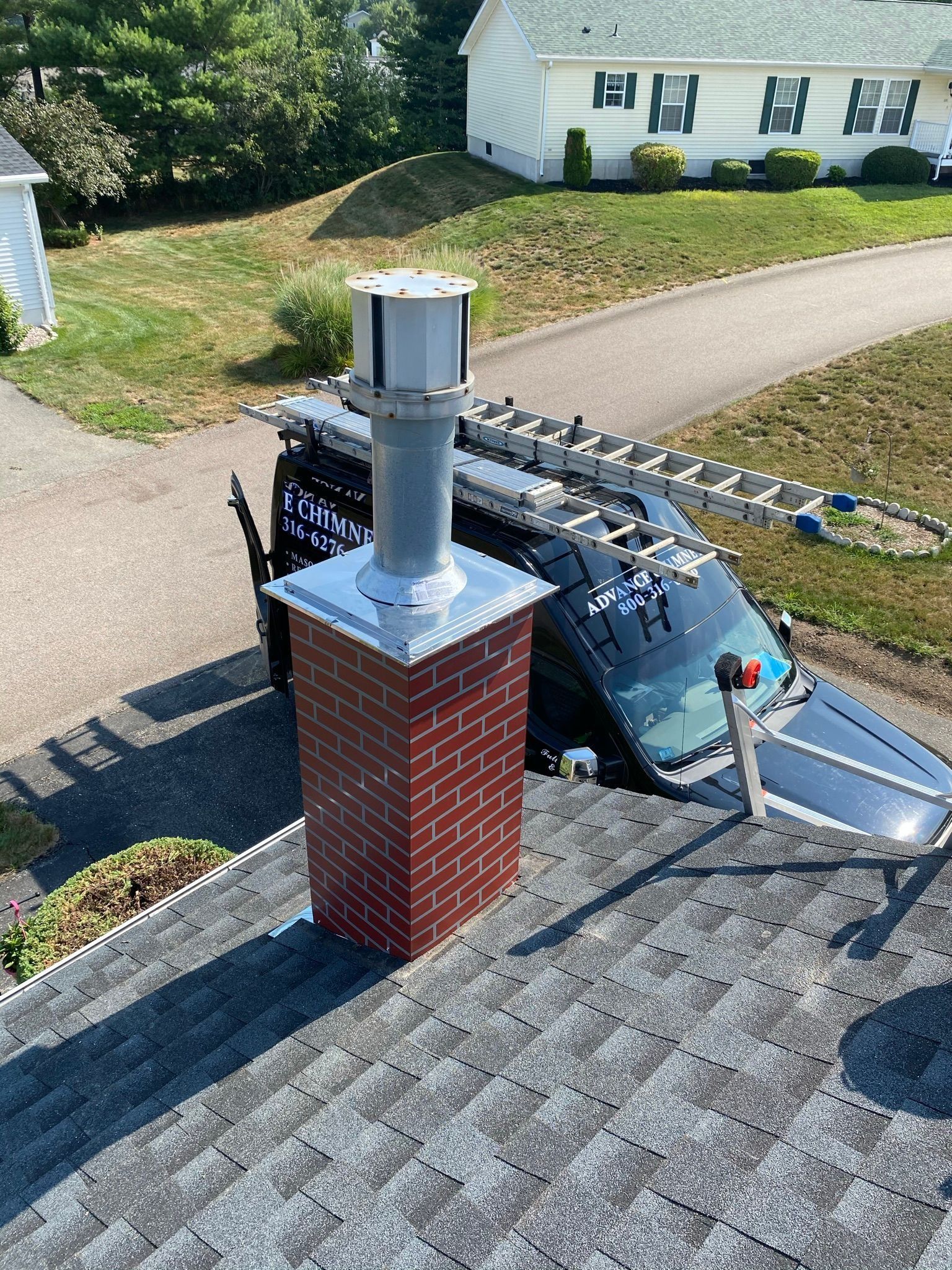 A brick chimney with a tall silver metal cap on a shingled roof, with a service van parked in the driveway below.
