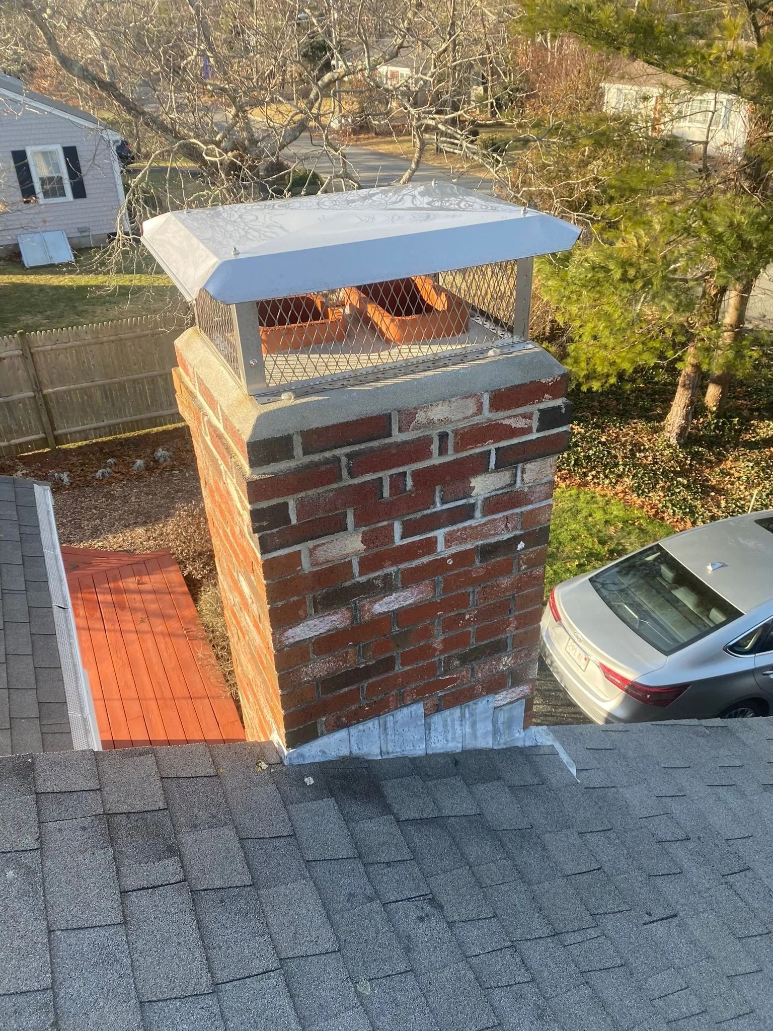 A brick chimney on a shingled roof with a metal cap, overlooking a yard with a parked car.