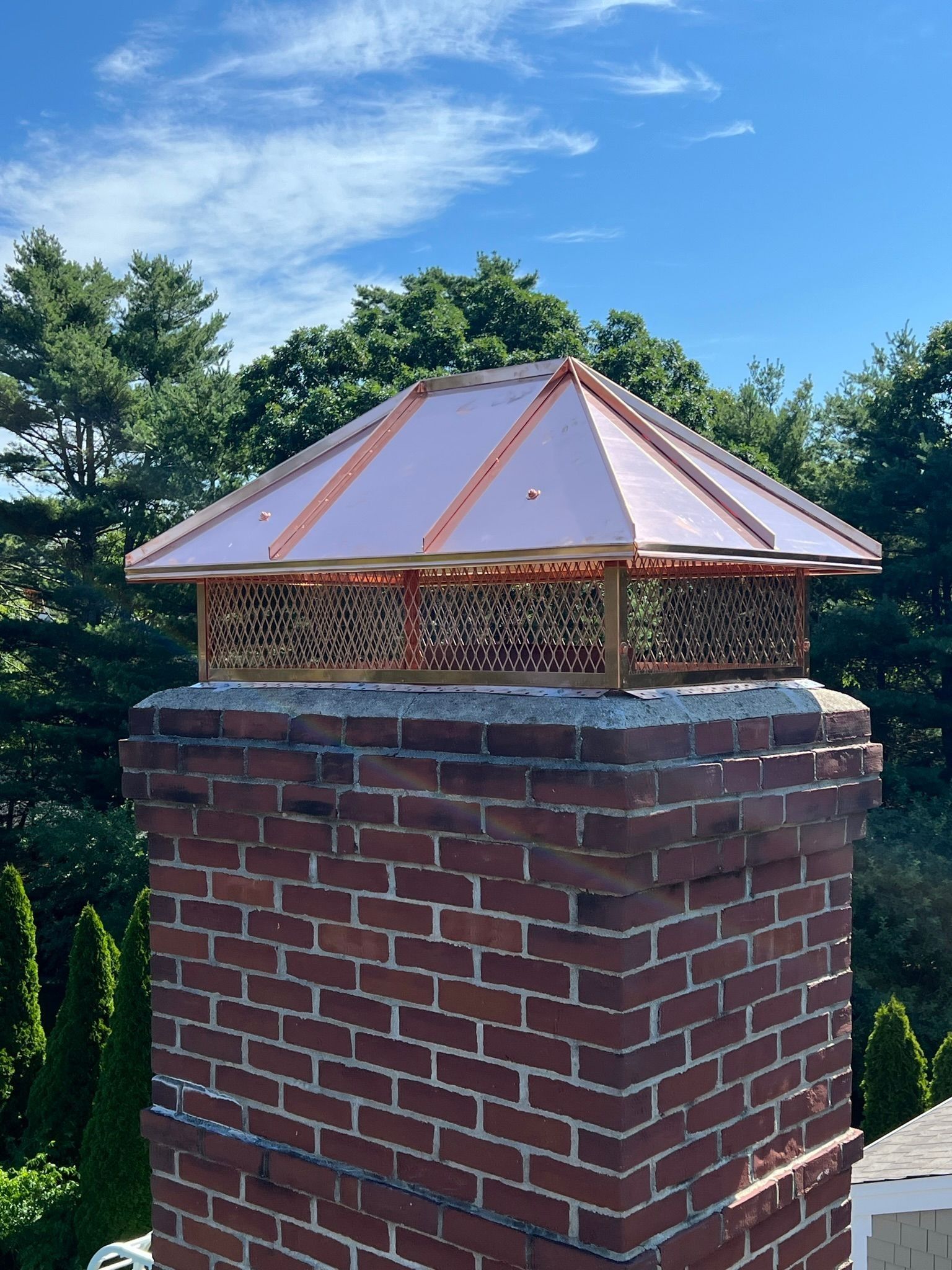 A brick chimney topped with an ornate copper-colored metal screen and a square, pyramid-shaped copper roof.