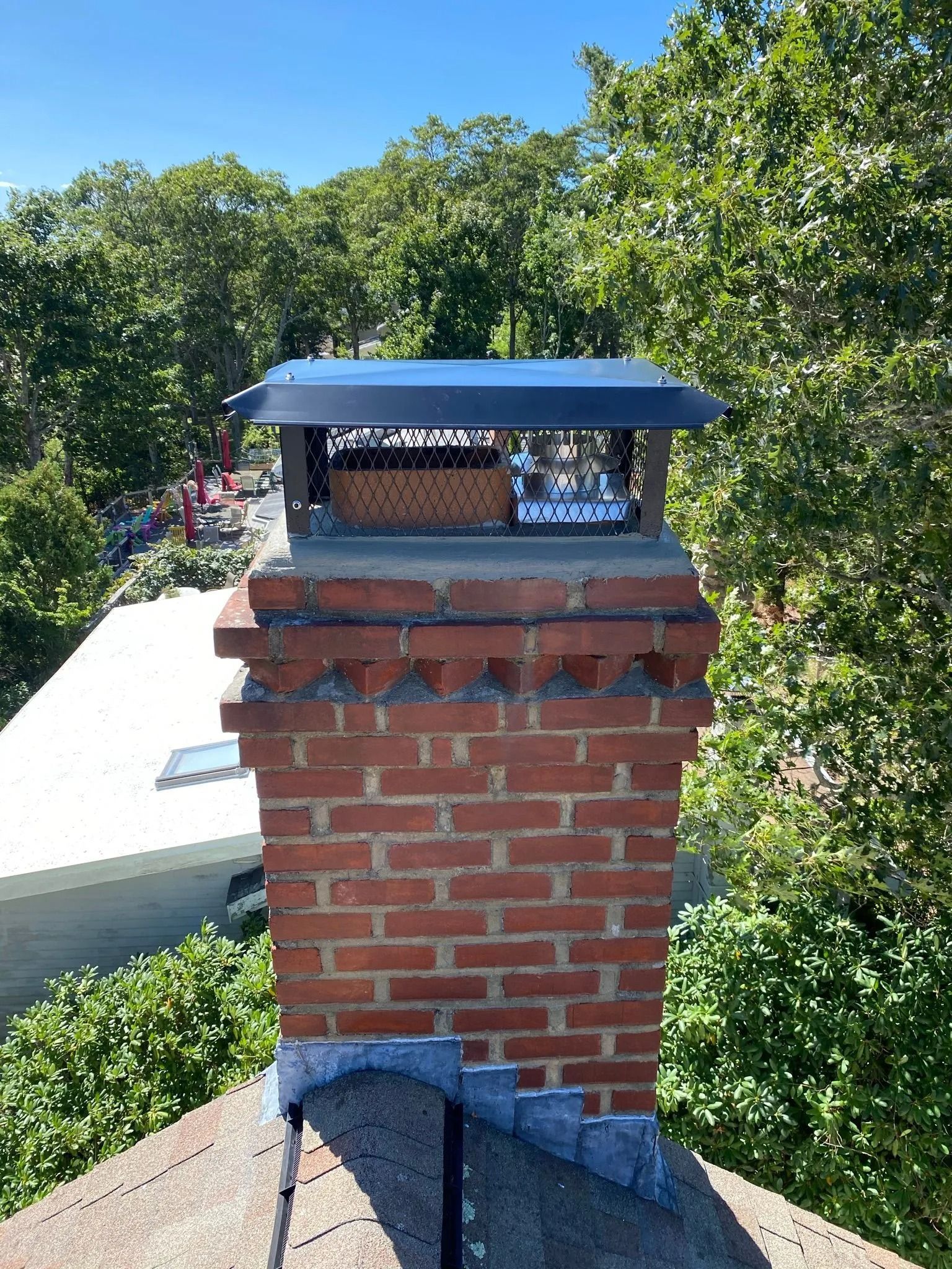 A brick chimney with a metal chimney cap on a roof, surrounded by green trees on a sunny day.