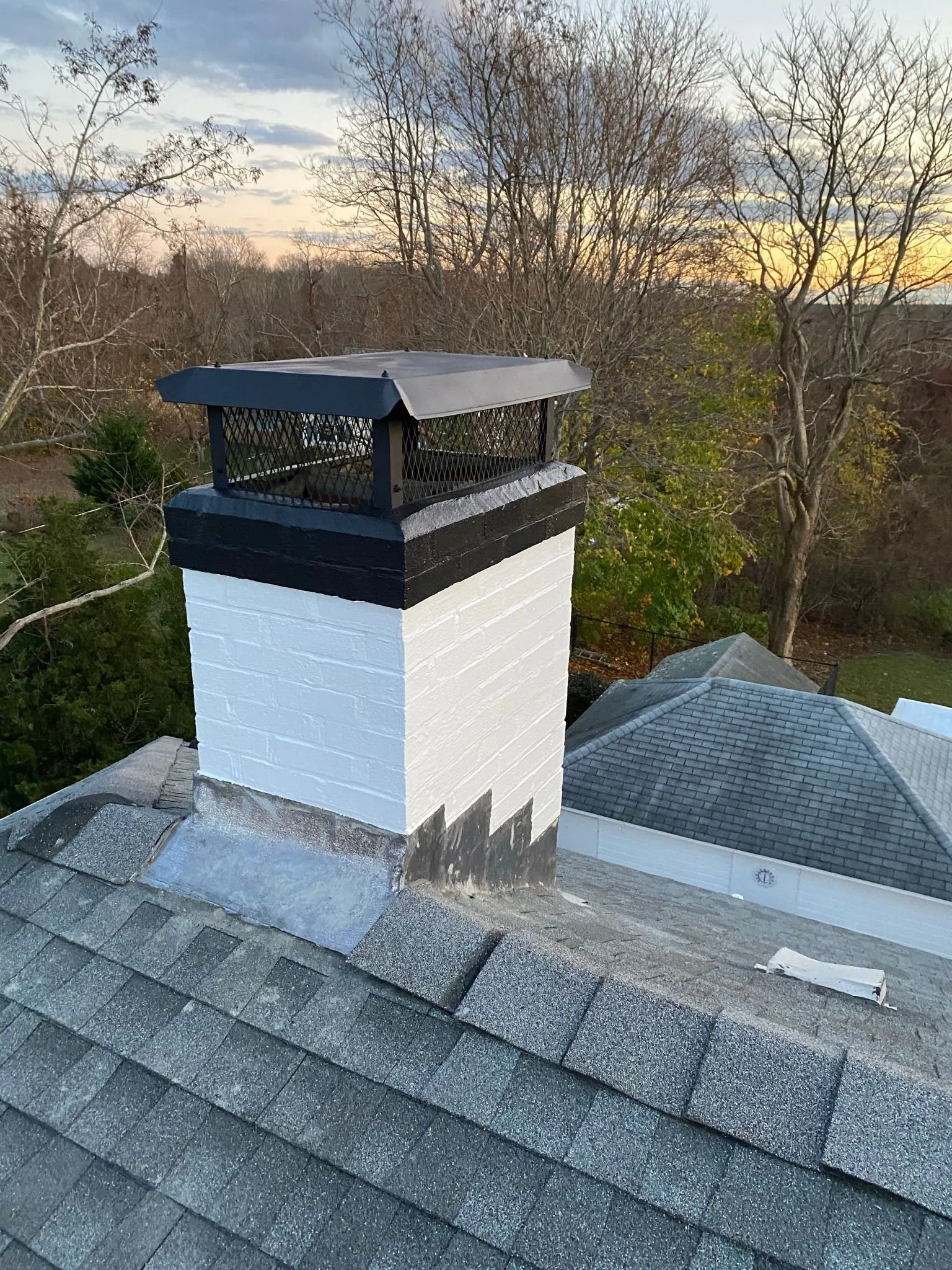 A painted white chimney with a black cap and base sits on a shingled roof against a background of trees at sunset.