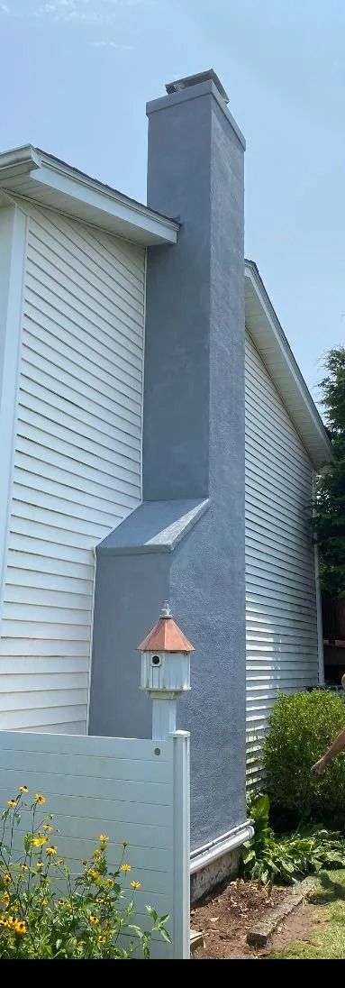 A gray stucco chimney attached to the white siding of a house, with a small decorative birdhouse on a post in the foreground.