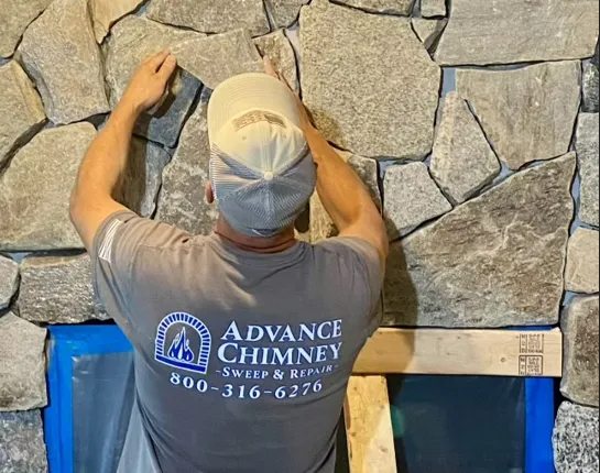 A worker in an Advance Chimney t-shirt installs natural stone veneer on a wall above a masked-off area.