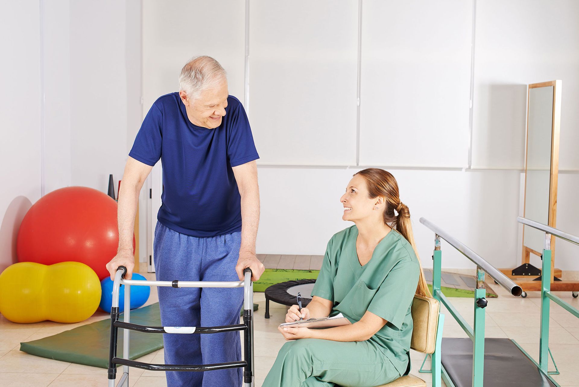 Man using a walker talks to a woman in scrubs in a rehabilitation gym.