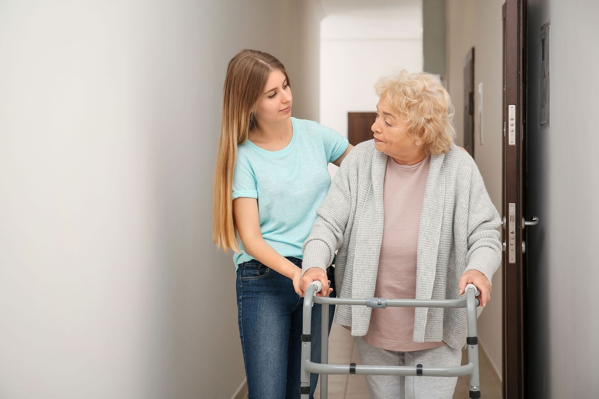 Young woman assists an older person with a walker down a hallway.