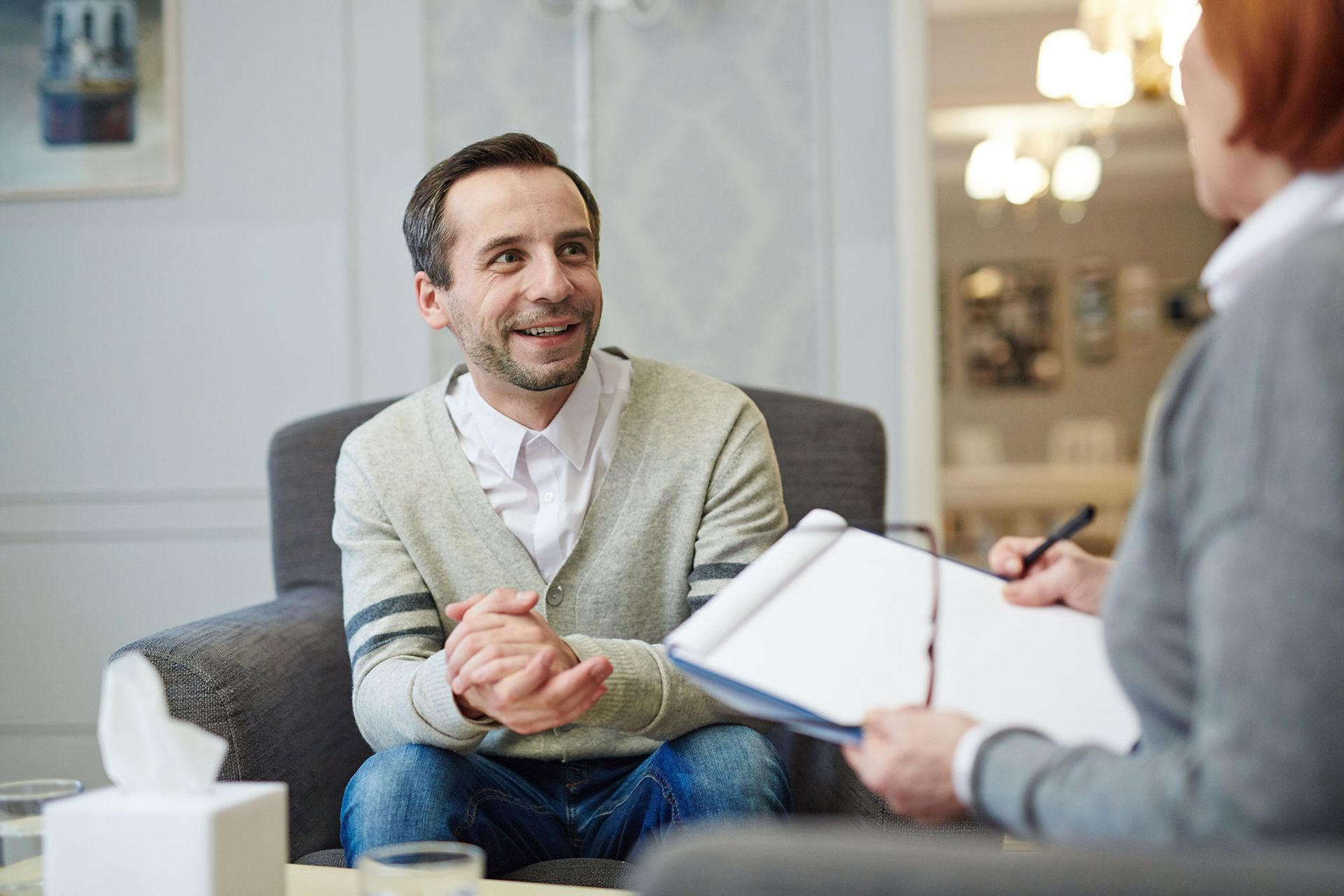 Man smiles in therapy, seated in armchair, facing a person with a clipboard; tissue box on the table.