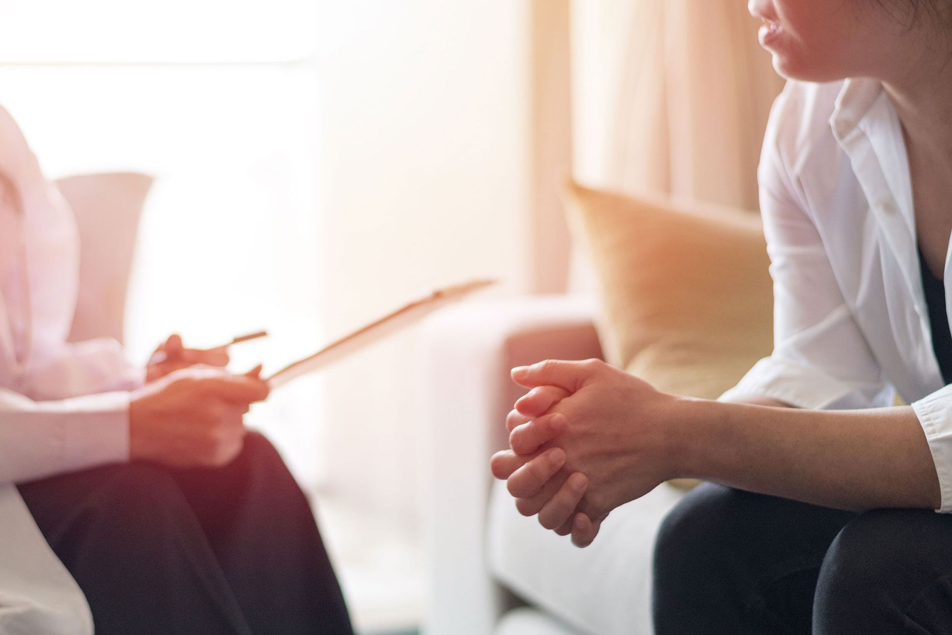 Person in session with therapist, listening attentively with hands clasped. Therapist holds clipboard in brightly lit room.