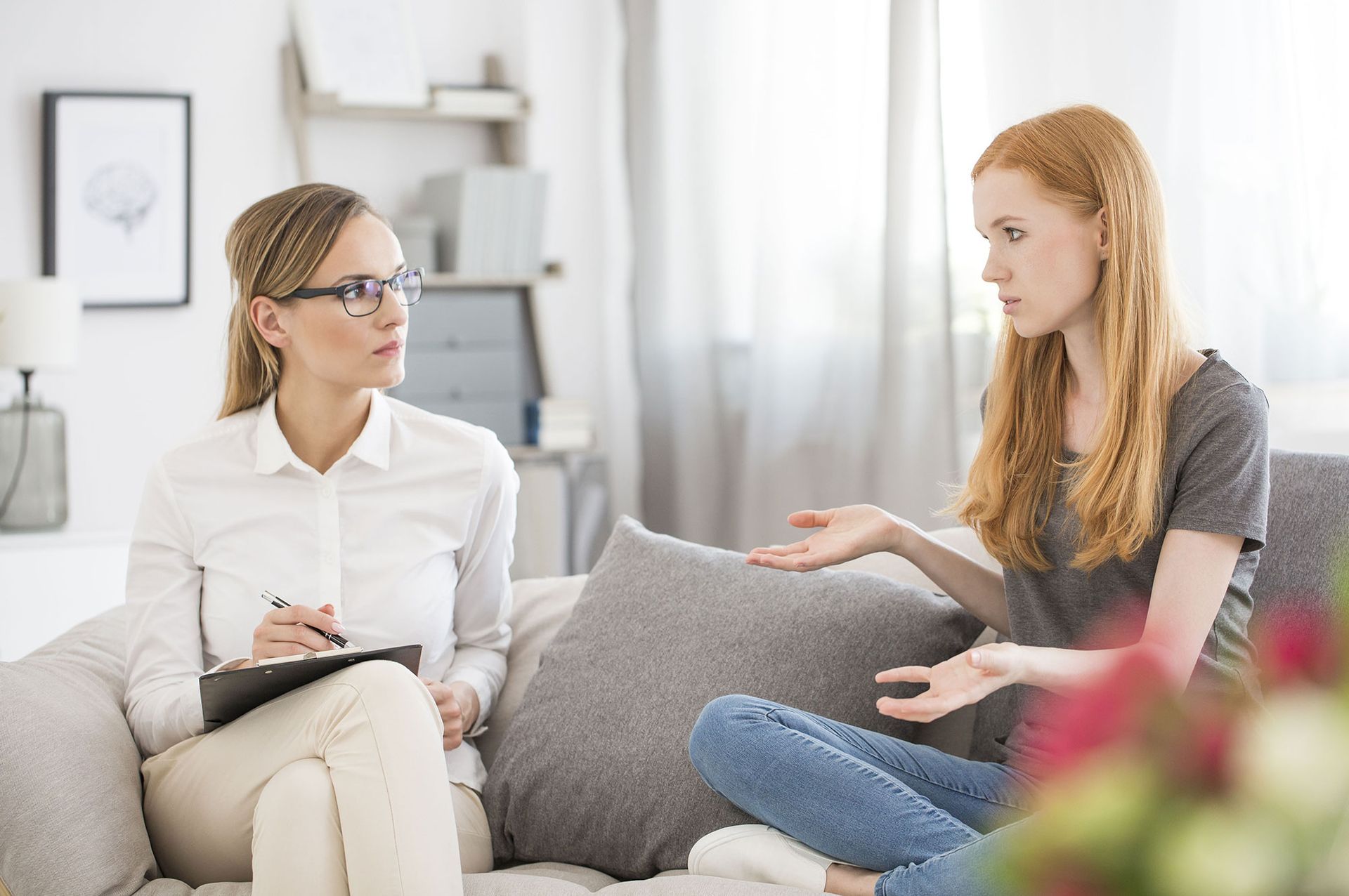 Woman in therapy session: therapist taking notes, patient gesturing, both on couch.