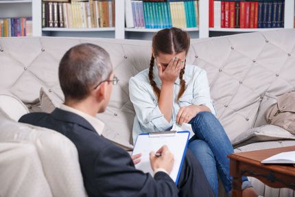 A person sits on a couch, crying, while another person writes on a clipboard.